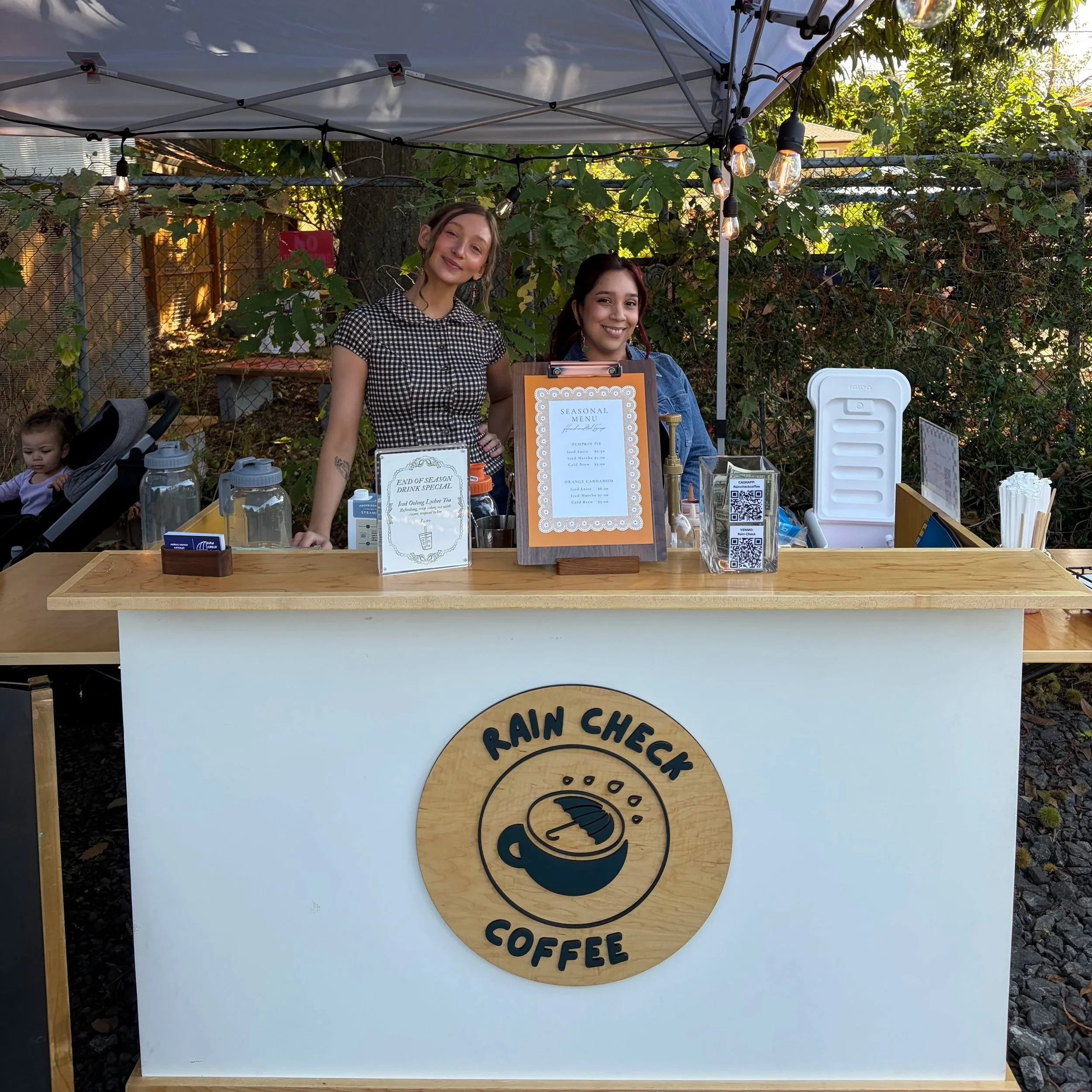 Two women standing behind a wooden table at an outdoor market stall with a sign that reads 'Seasonal Menu' and a coffee logo, surrounded by greenery and string lights overhead.