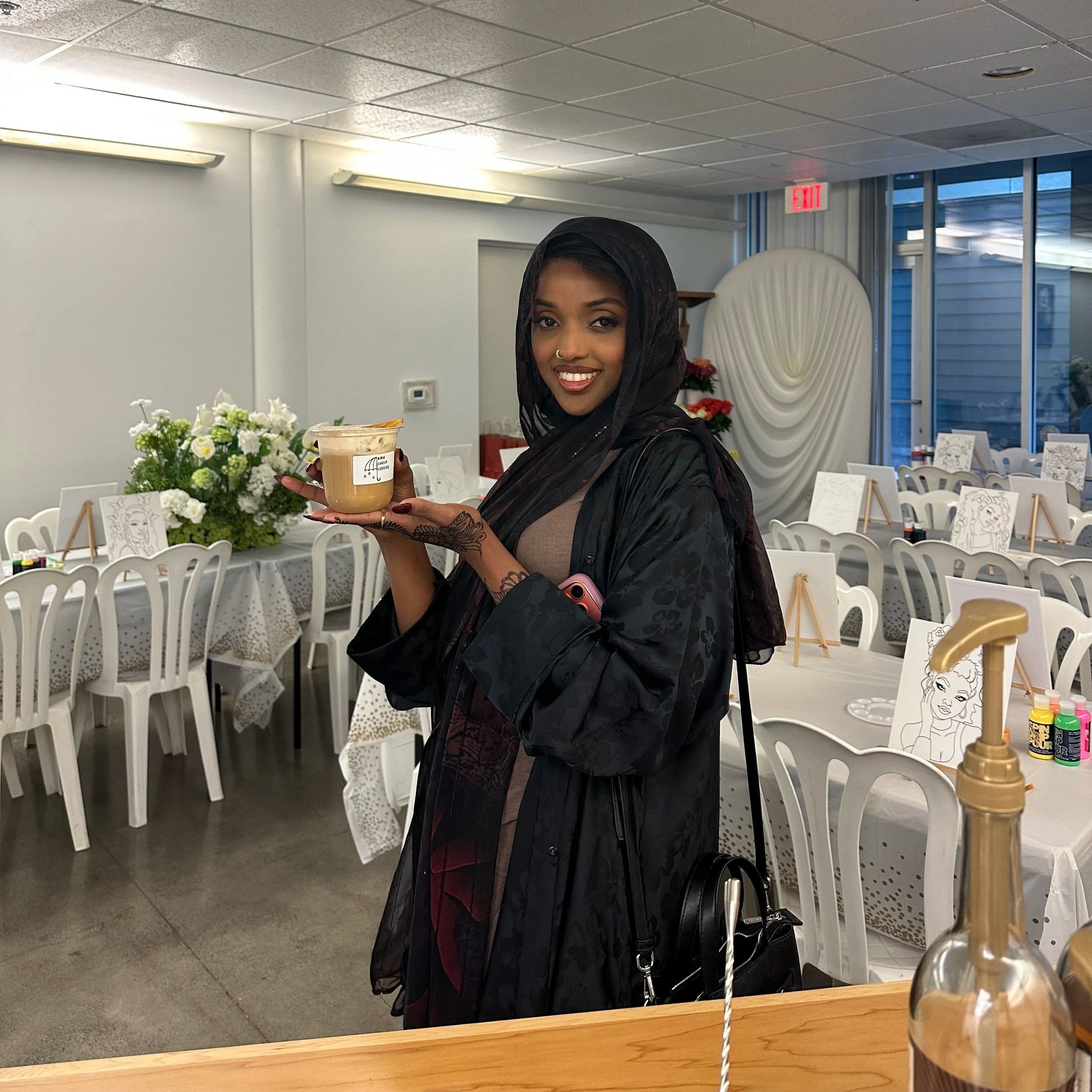 A woman smiling and holding a plastic cup of iced coffee in a decorated indoor setting with white chairs, tables, floral arrangements, and drawing easels.