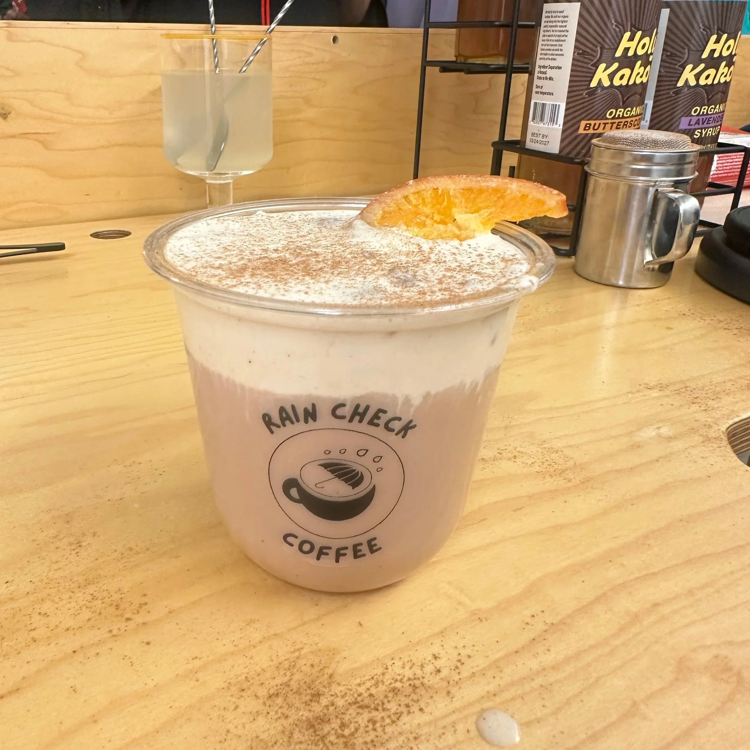 A transparent plastic cup with a whipped cream topping dusted with cinnamon, a piece of caramelized sugar or tuile, a glass of water with two metal straws, a metal sugar shaker, and two boxes of organic syrup on a wooden surface in a cafe.