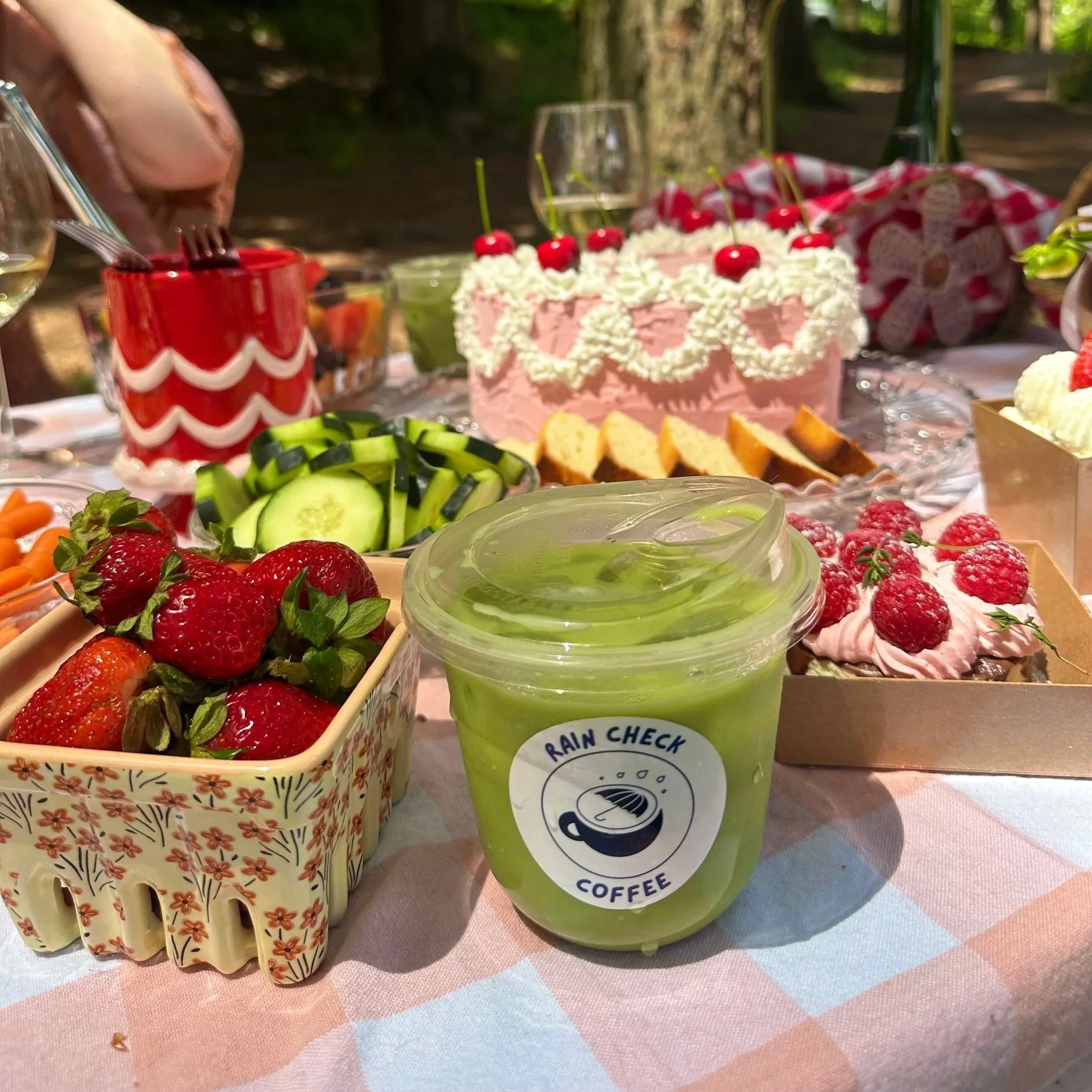 A picnic table set with a variety of food and drinks, including a pink frosted cake with cherries on top, strawberries, sliced cucumbers, and a green smoothie labeled "Rain Check Coffee," in a forested outdoor setting.