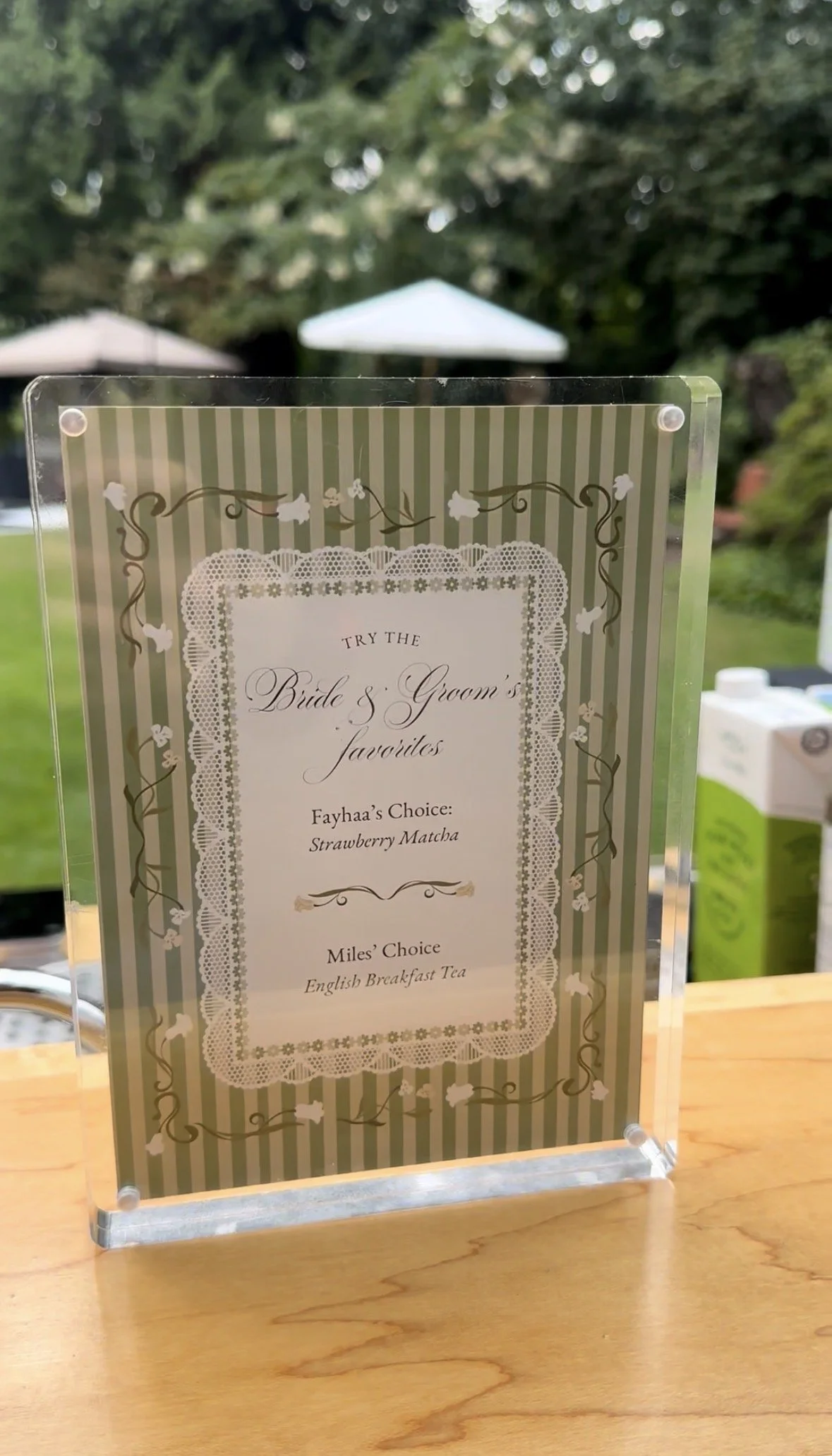 A sign at a wedding reception with a list of bride and groom's favorite drinks, including strawberry matcha and English breakfast tea, in an outdoor garden setting with umbrellas in the background.