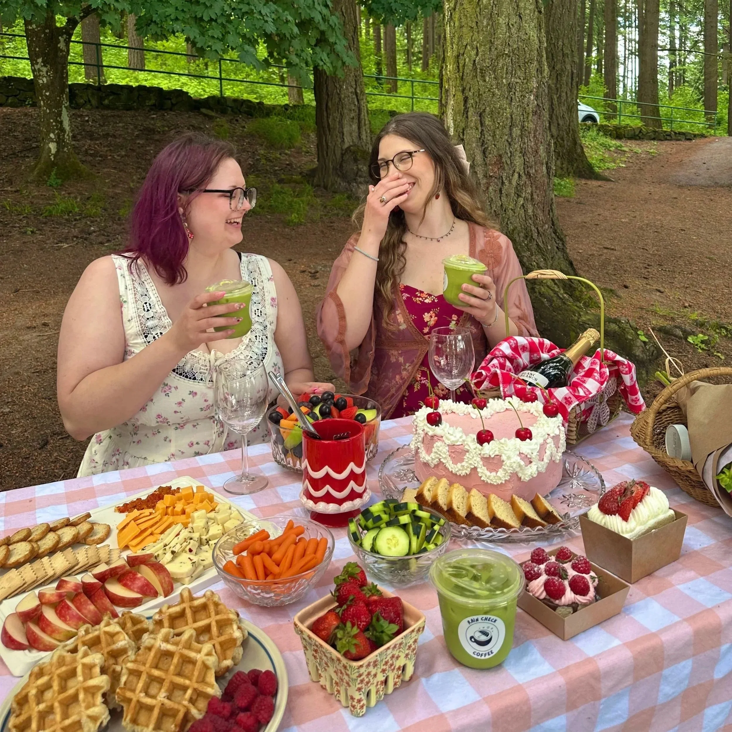 Two women sitting outdoors at a picnic table filled with food, including fruit, waffles, and a cake with cherries. One woman has glasses and reddish-pink hair, the other has glasses and long brown hair. They are smiling and holding green drinks. The background shows trees and a forested area.