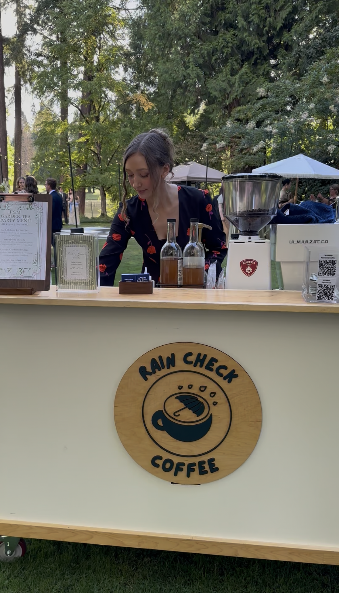 A woman working at a coffee stand outdoors with trees and people in the background. The stand has a sign that reads "Rain Check Coffee" and features a logo of a coffee cup with an umbrella.