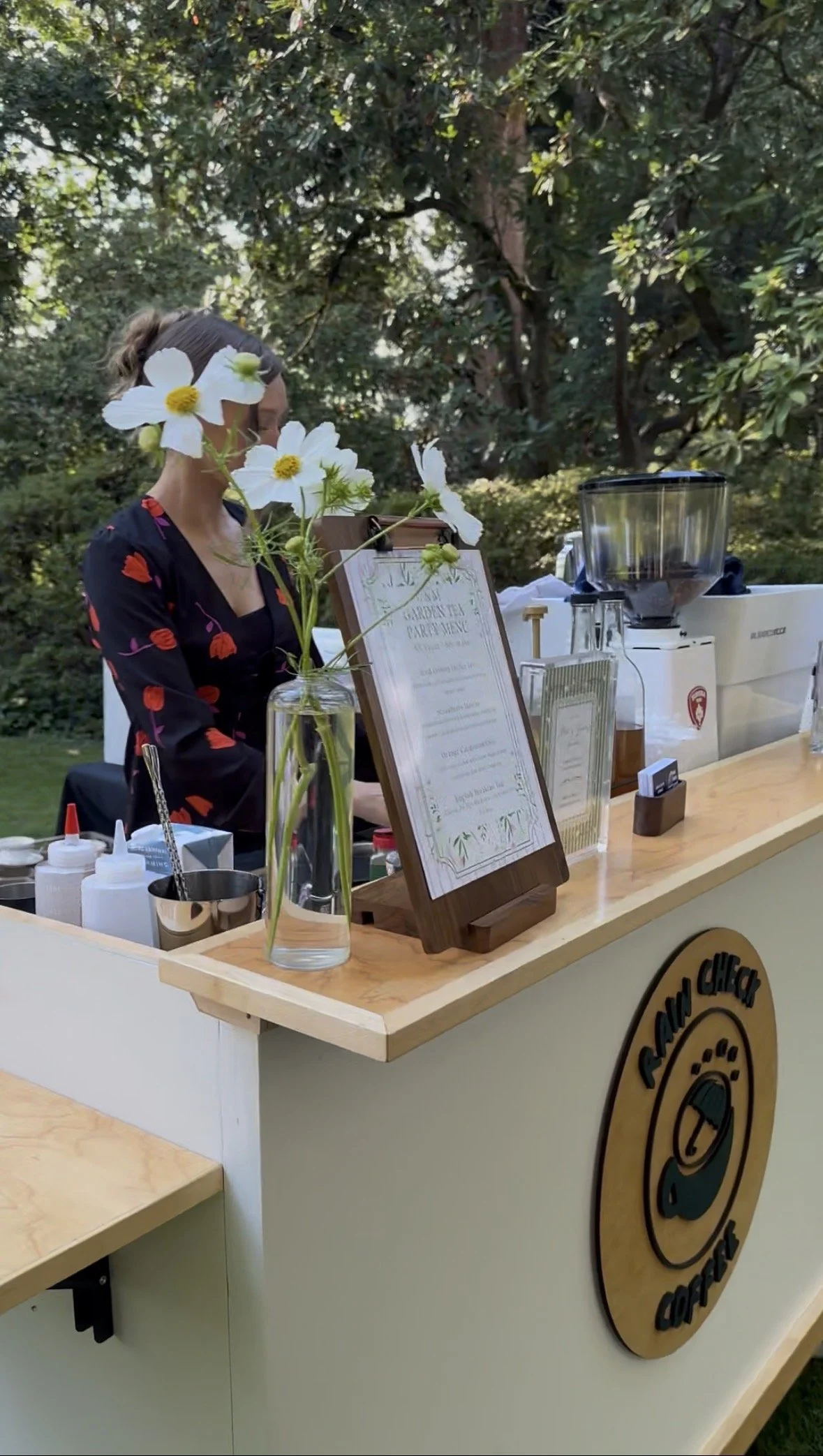 A woman working at a coffee stand outdoors, with trees in the background, a menu on a stand, and various supplies on the counter.