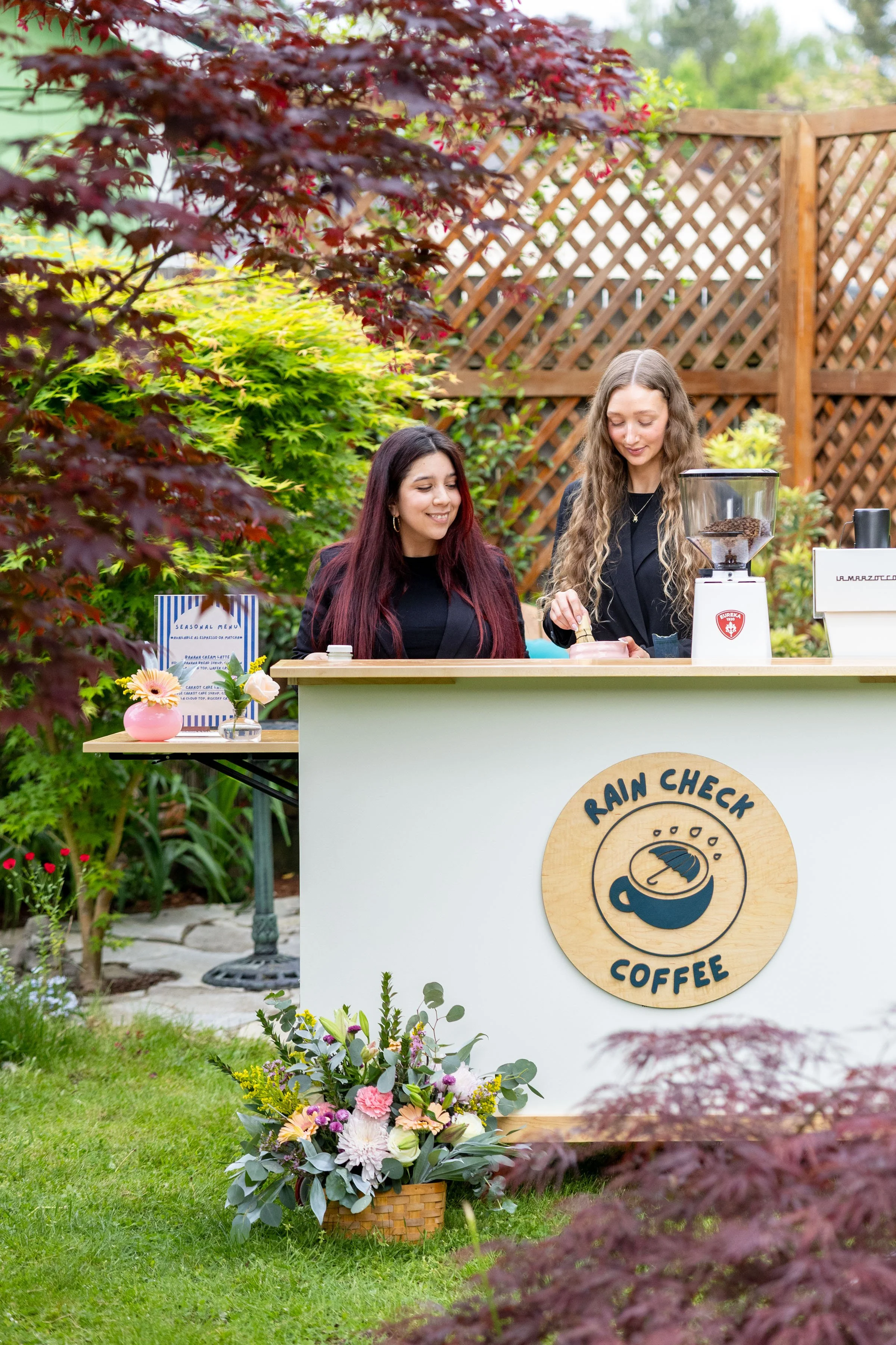 Two women serve coffee at an outdoor coffee stand surrounded by green and purple foliage. The stand has a sign with a coffee cup logo and the words "Rain Check Coffee." There are flowers in a basket in the foreground.