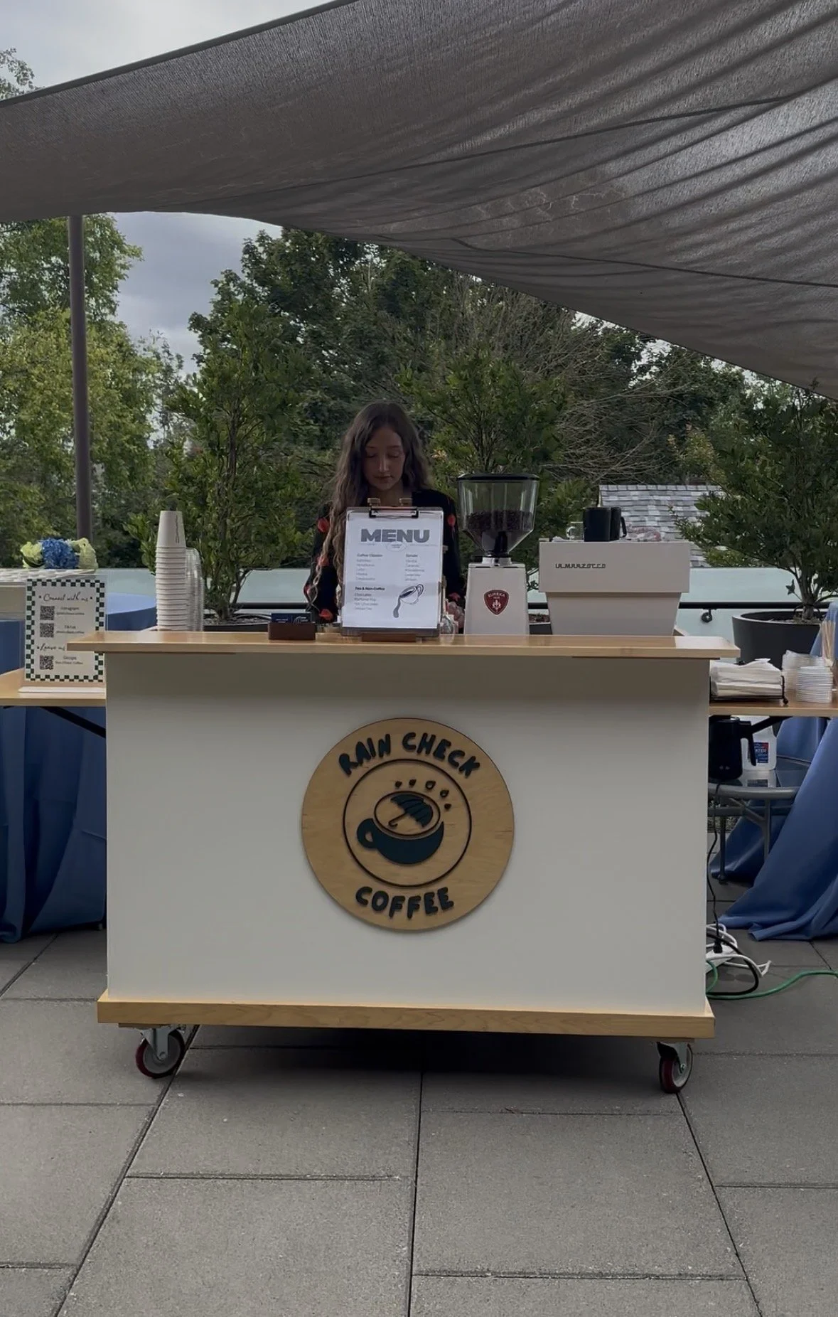A girl working at a mobile coffee stand called 'Rain Check Coffee' outdoors with trees in the background. The stand has a menu, a coffee grinder, and disposable cups.