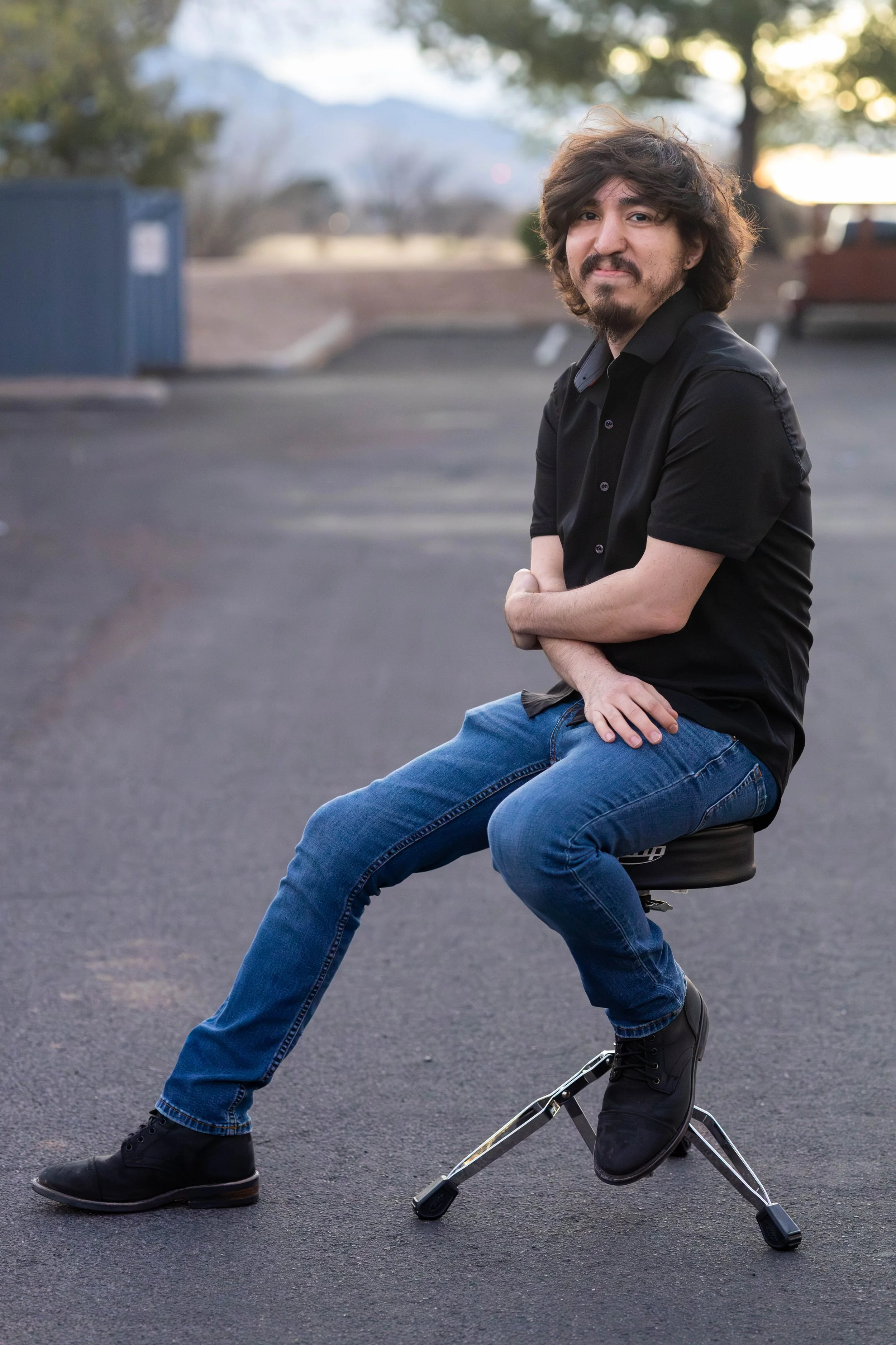 A man with curly hair and beard, wearing a black short-sleeve shirt and blue jeans, sitting on a stool with unusual metallic legs in a parking lot during sunset.