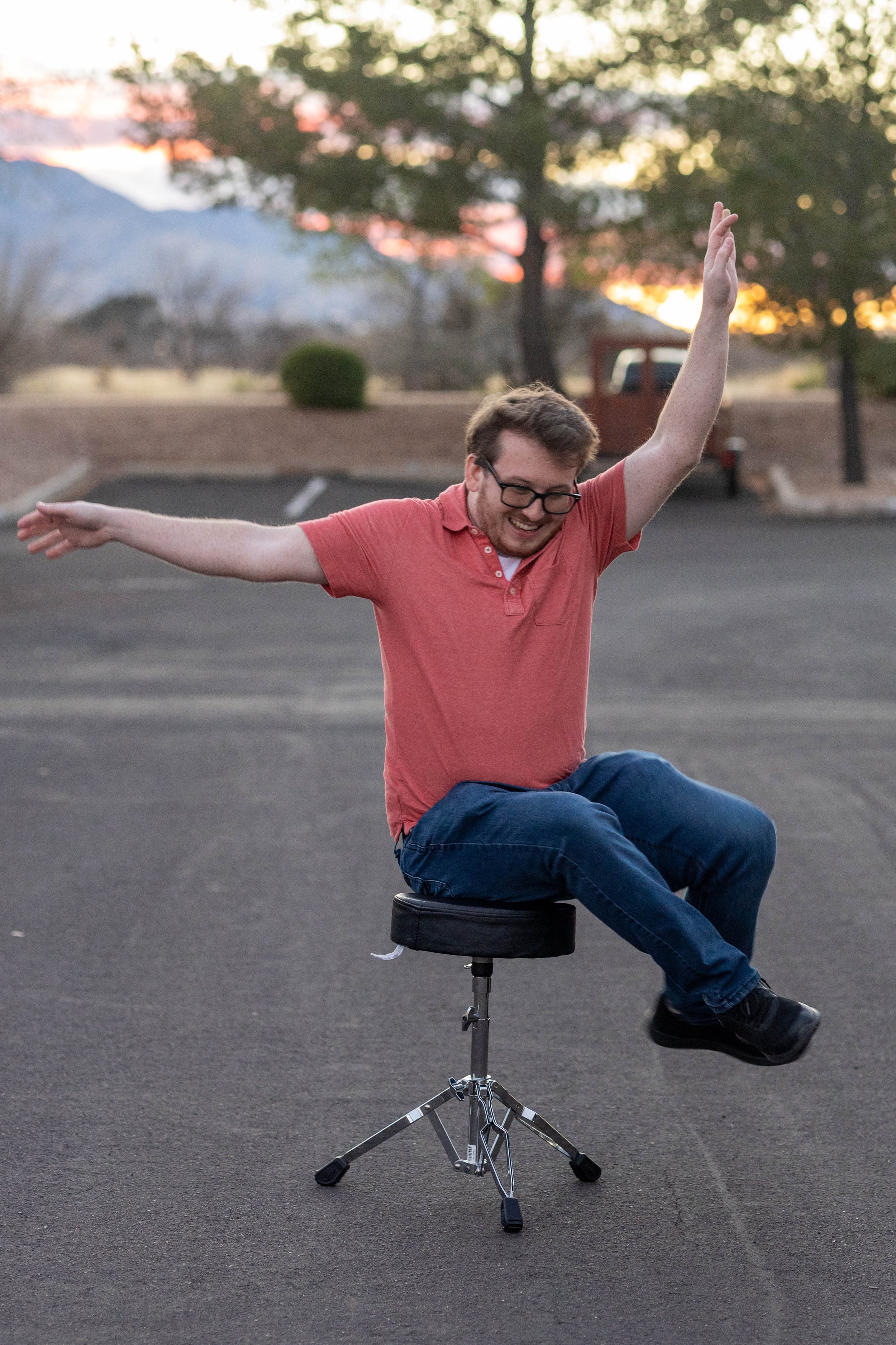 A man with glasses, wearing a salmon polo shirt and jeans, sitting on a drummer's stool in an empty parking lot at sunset, smiling with his arms outstretched.