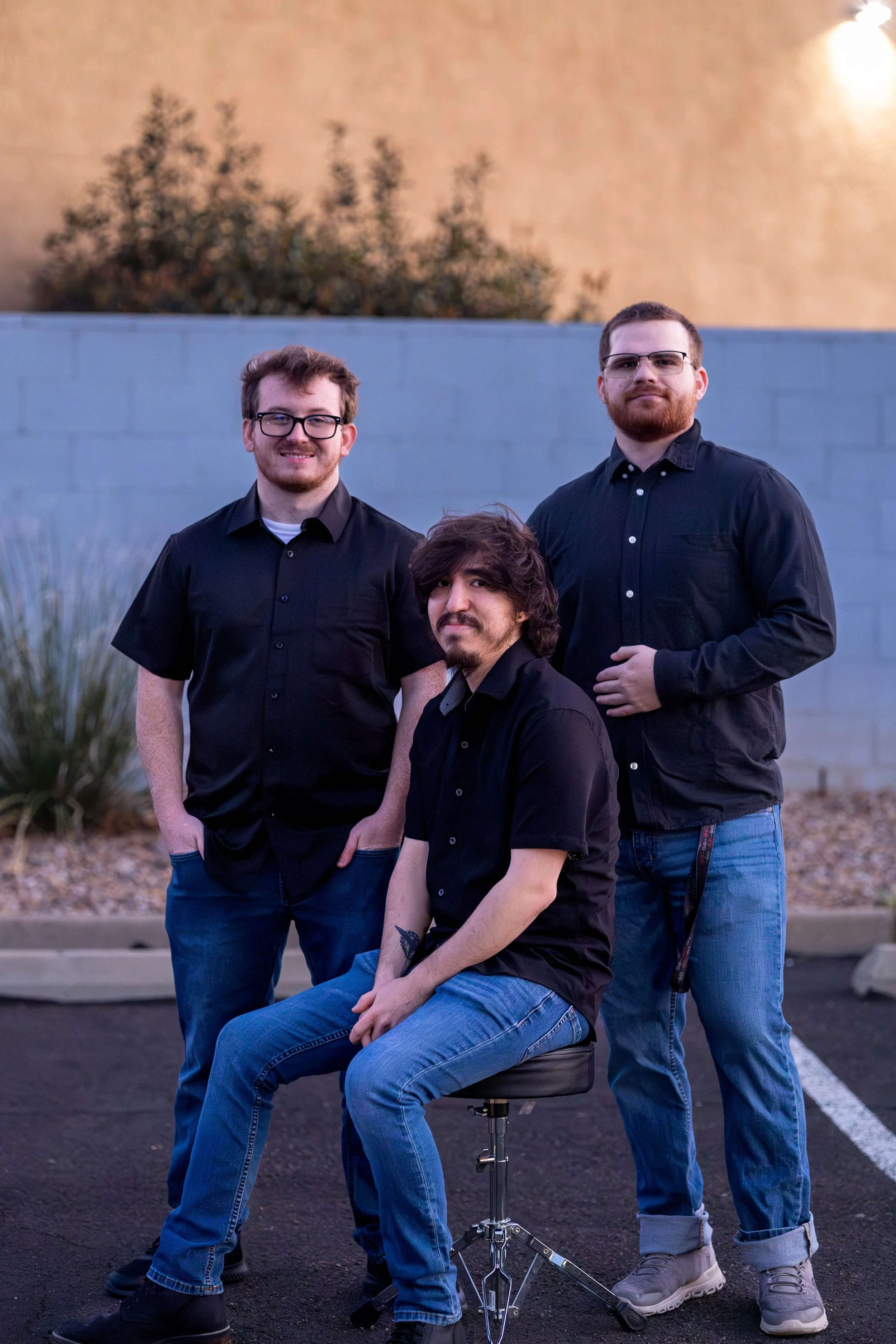 Three men in black shirts and blue jeans pose outdoors against a wall, with one sitting on a black stool in front of the other two.