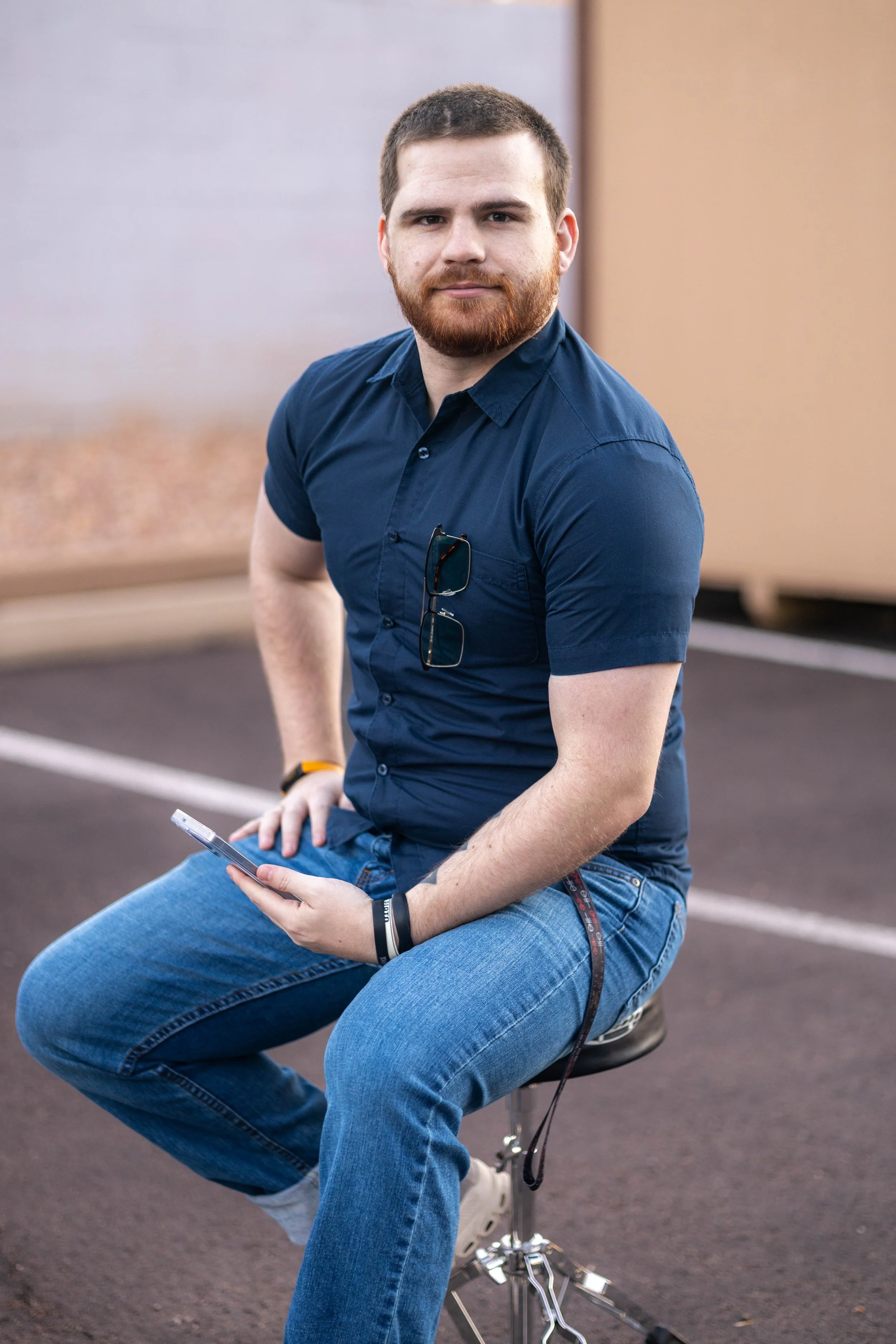 A young man with short brown hair, beard, wearing a navy blue shirt with sunglasses on the chest, sitting on a stool outside holding a smartphone, with a pinkish-orange wall in the background.