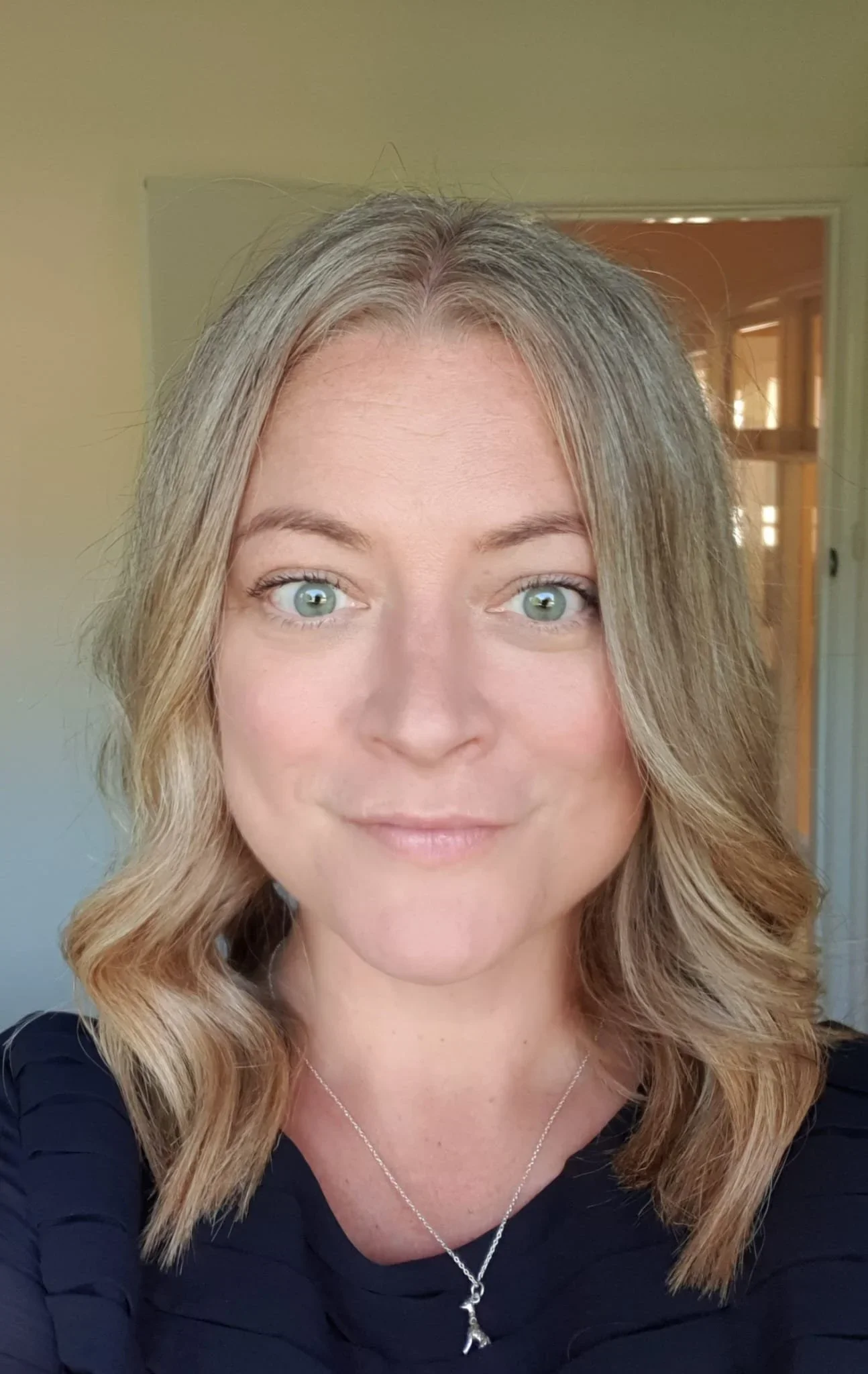 A woman with wavy, blonde hair and light green eyes, wearing a black top and a silver necklace, smiling in front of a beige wall and an open doorway with warm lighting.