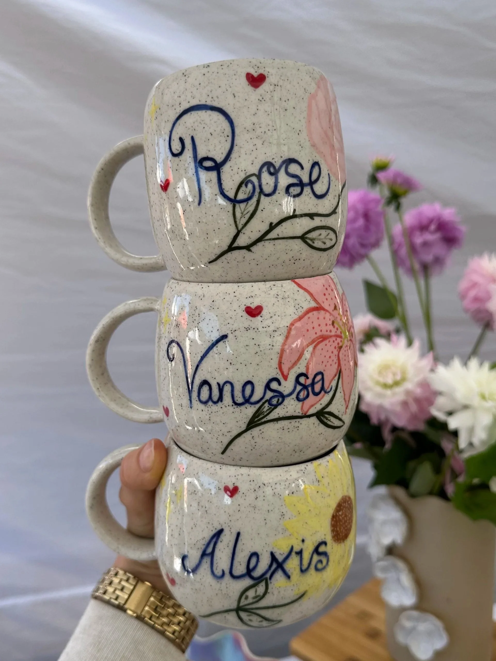 Stack of ceramic cups with hand-painted names and designs, with a person holding them wearing a gold watch and a light-colored sweater, flowers in the background.