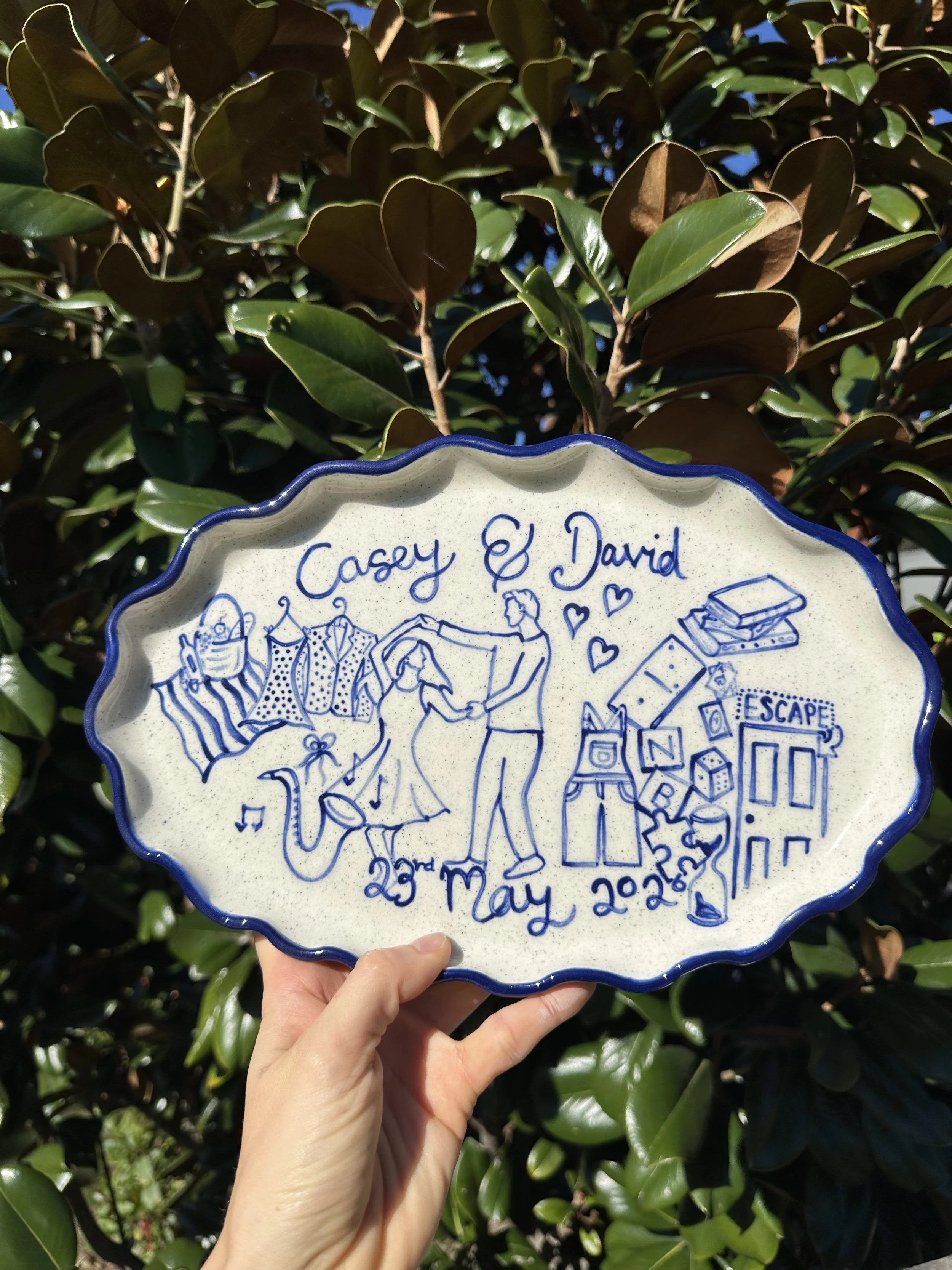 A ceramic plate with blue border and blue handwritten text reading "Casey & David" and the date "23rd May 2023". The plate has illustrations of a couple, a dog, books, and hearts, held up in front of a leafy bush.