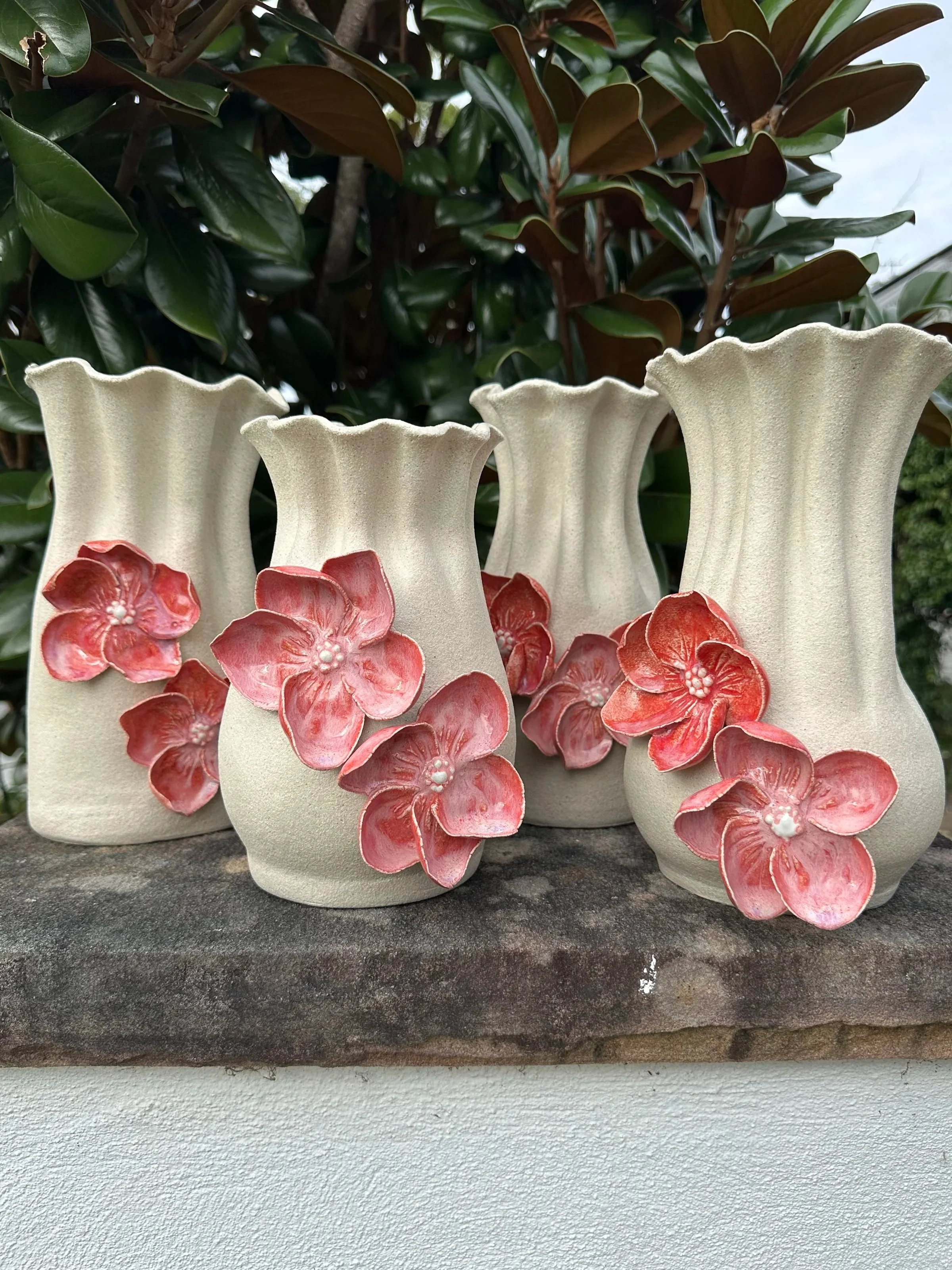 Three cream-colored ceramic vases with pink and red floral designs, placed on a stone surface with greenery in the background.