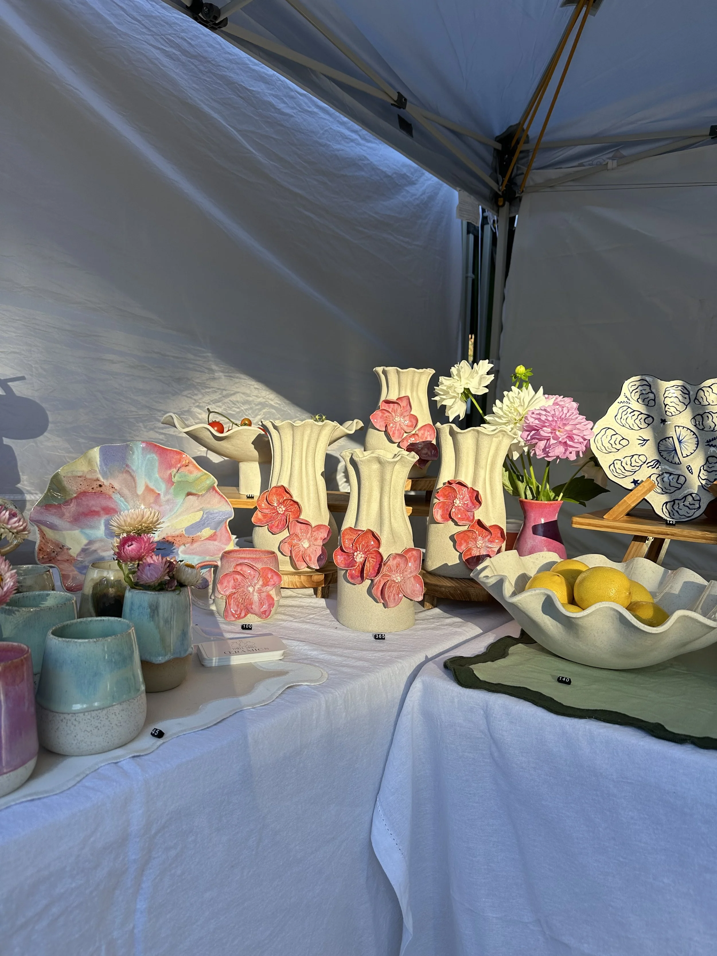 Display of colorful ceramics, vases, bowls, and flowers on a table at an outdoor market or craft fair under a tent.