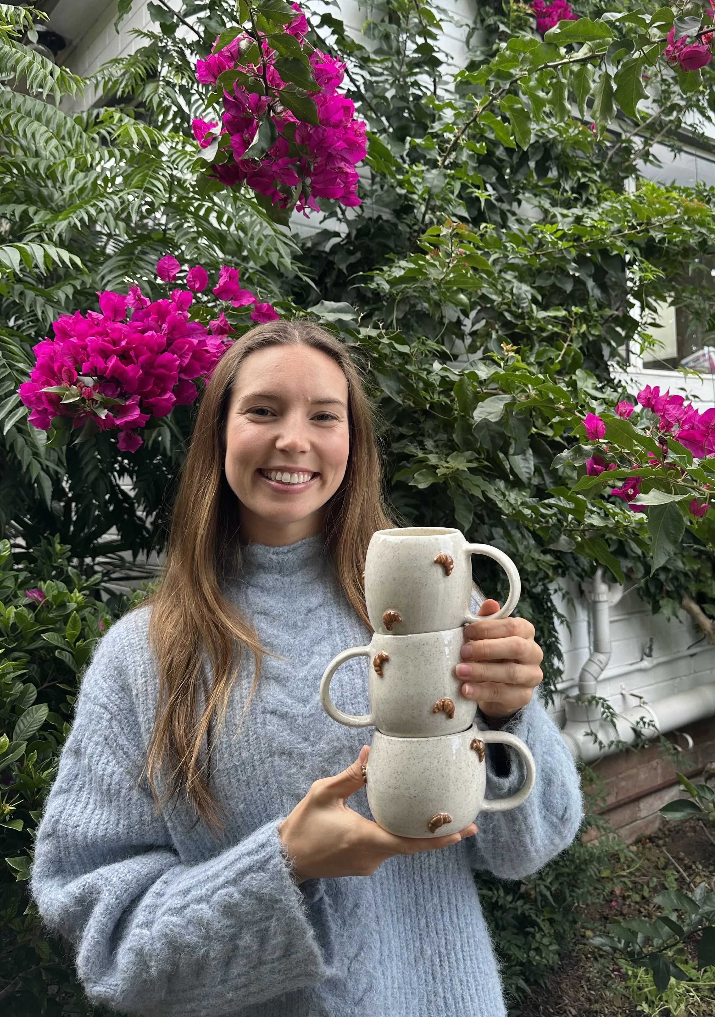 A young woman smiling and holding a stack of four white ceramic mugs with small brown decorations, standing outdoors in front of green plants with pink flowers.