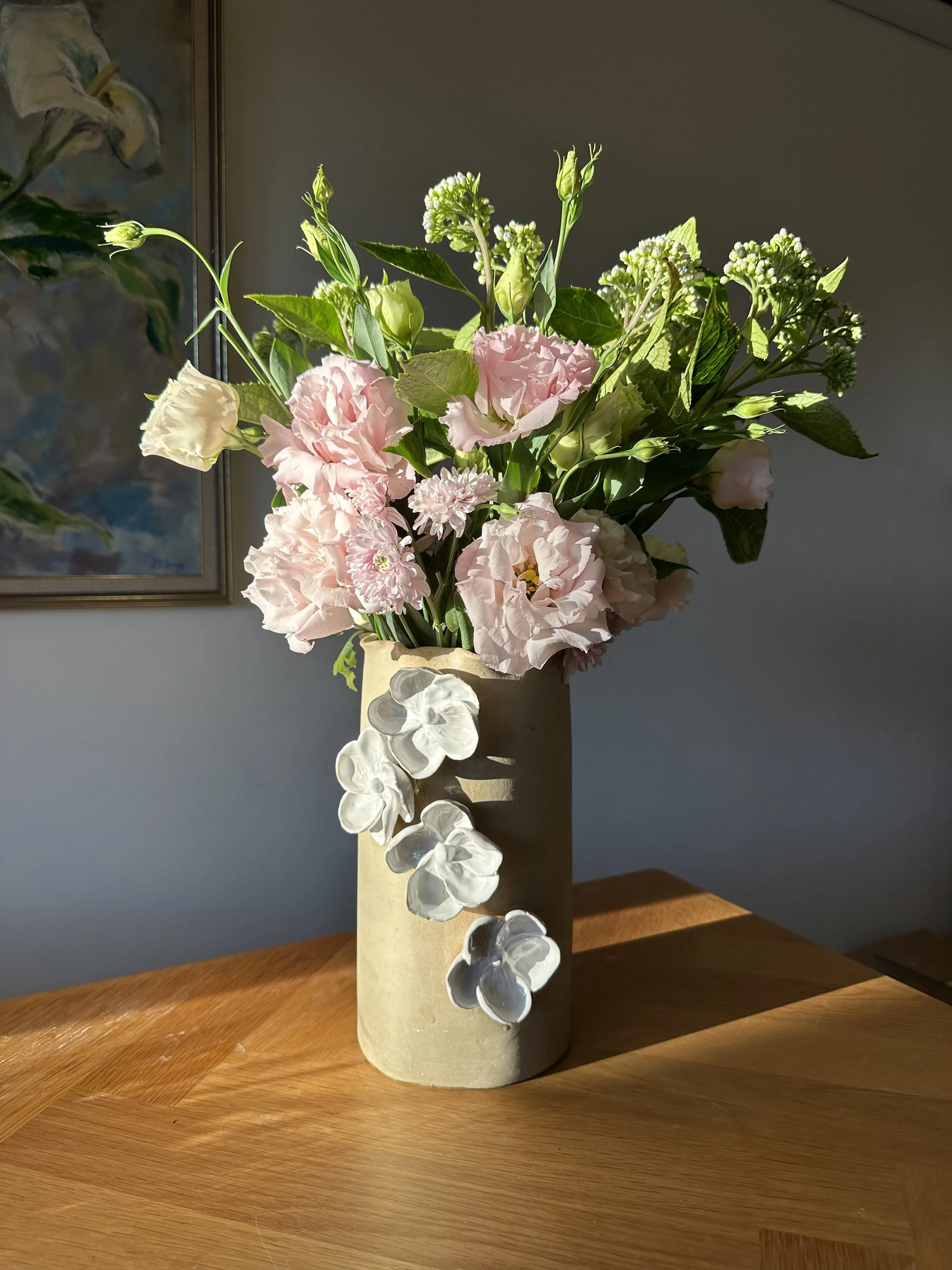 A beige ceramic vase with white ceramic flowers arranged with light pink and white flowers, and green leaves, placed on a wooden surface with a painting in the background.