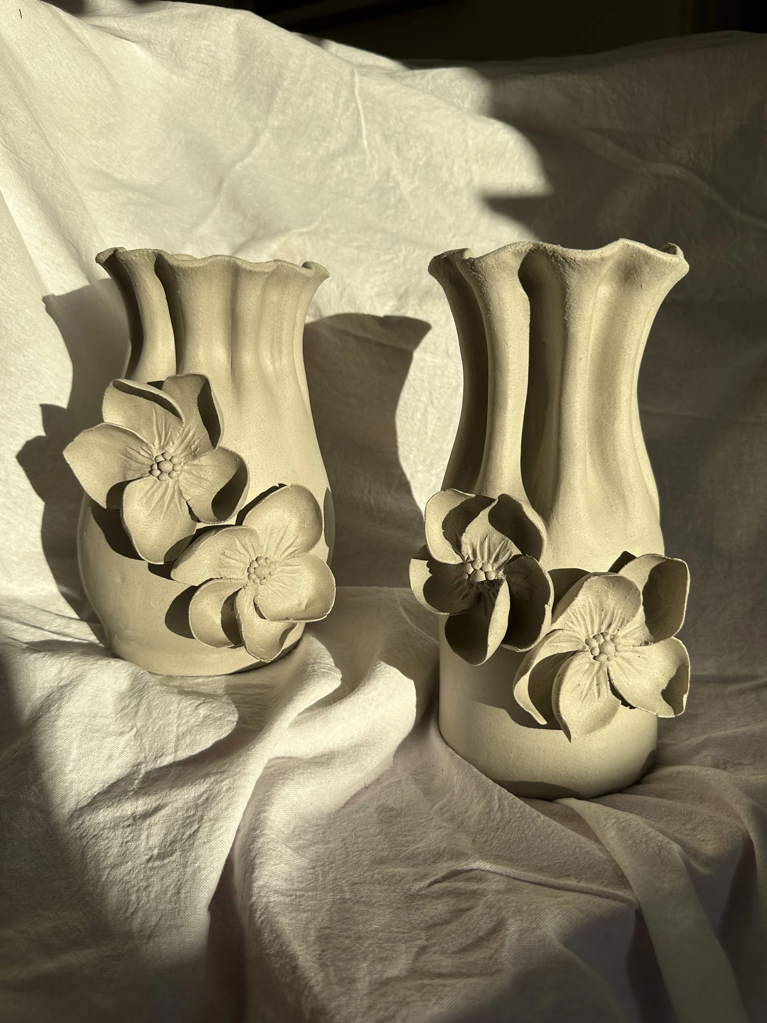 Two beige ceramic vases with floral and floral-like designs, placed on a white fabric background, with sunlight casting shadows.