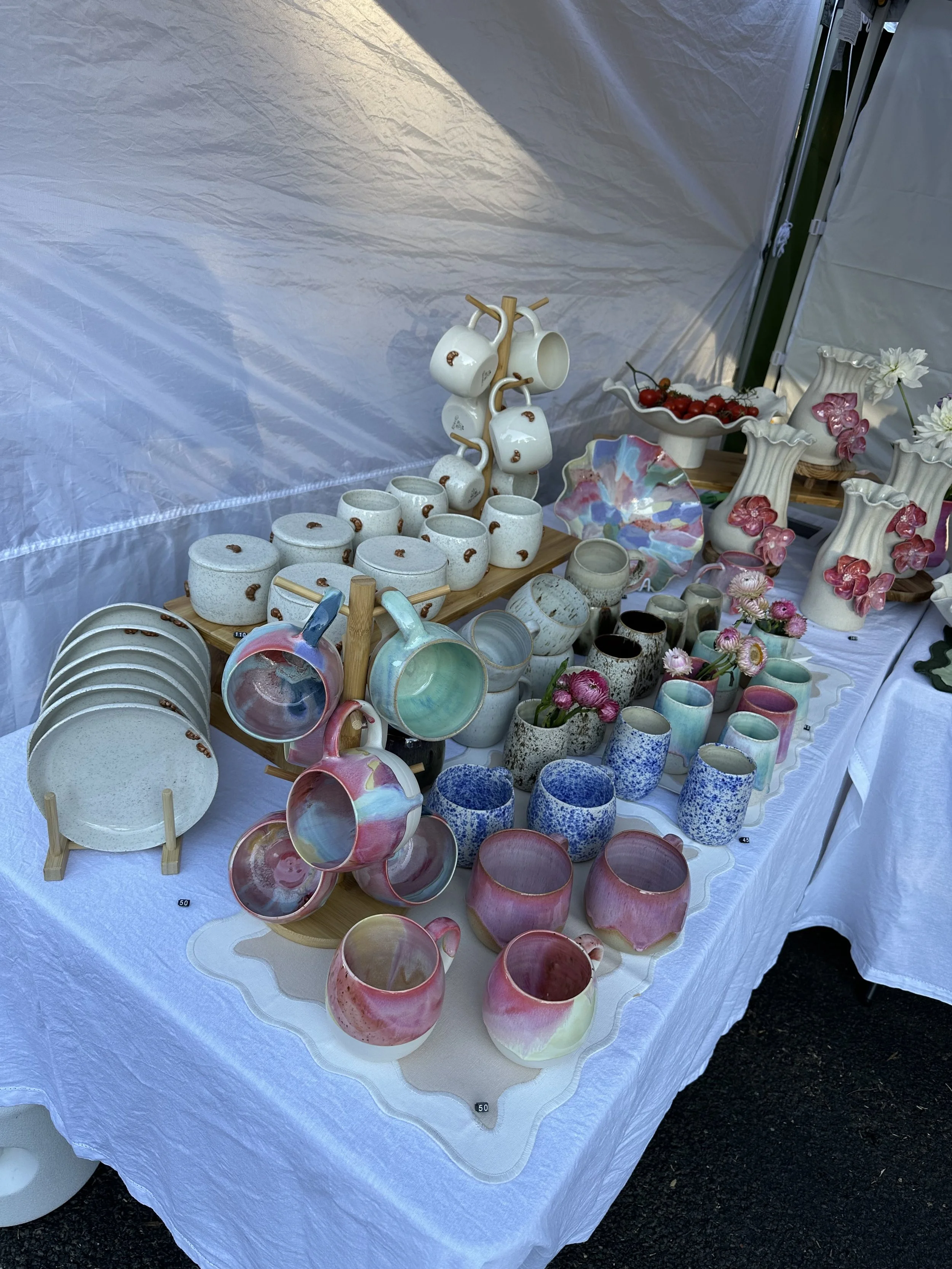 A table displaying various handmade ceramic pottery items, including cups, bowls, vases, and decorative plates, all with colorful and pastel glazes at an outdoor market stall.