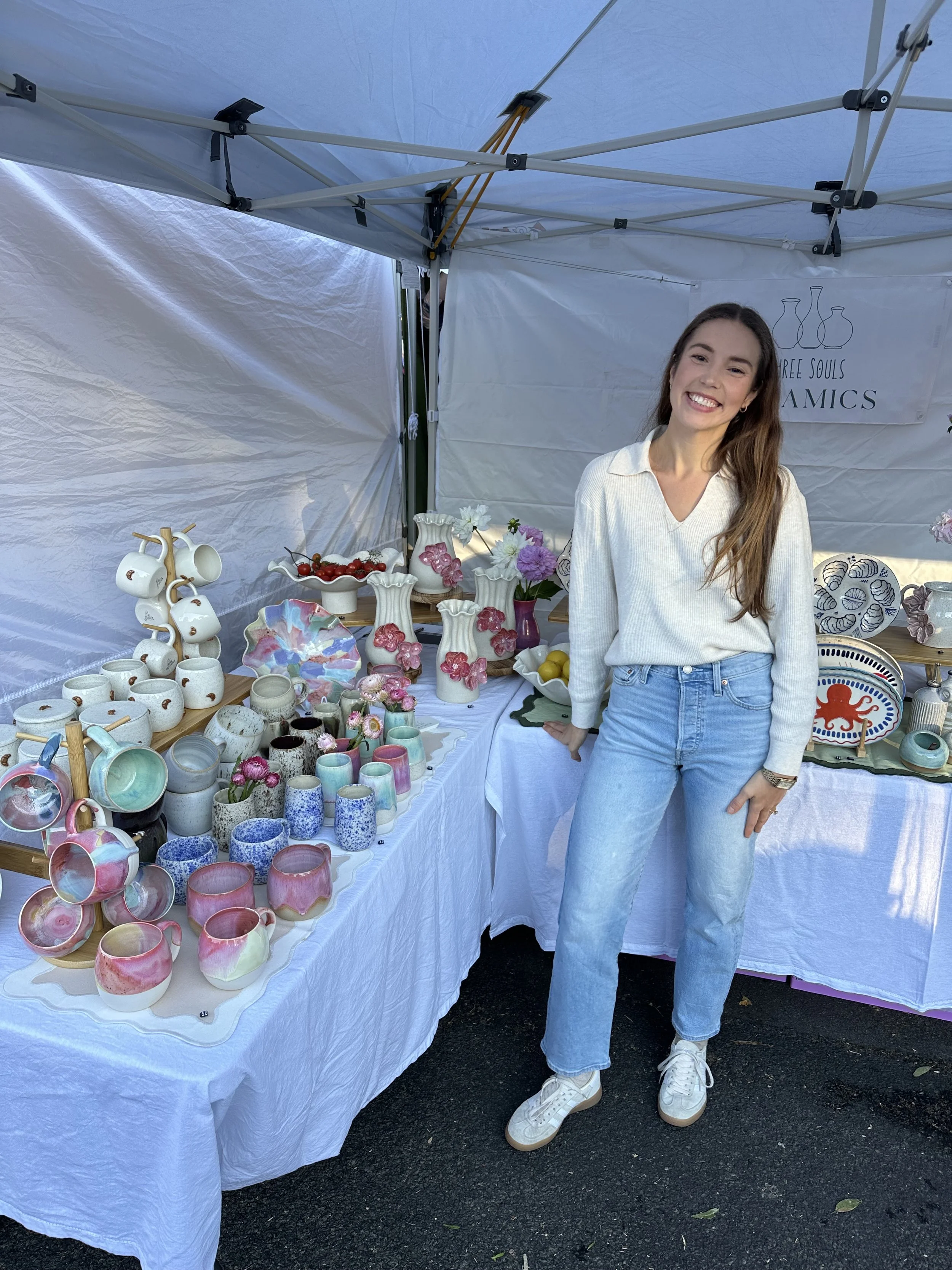 A woman with long brown hair, wearing a white sweater and light blue jeans, smiling and standing next to a table of ceramic pottery and decorative items at an outdoor market stall.