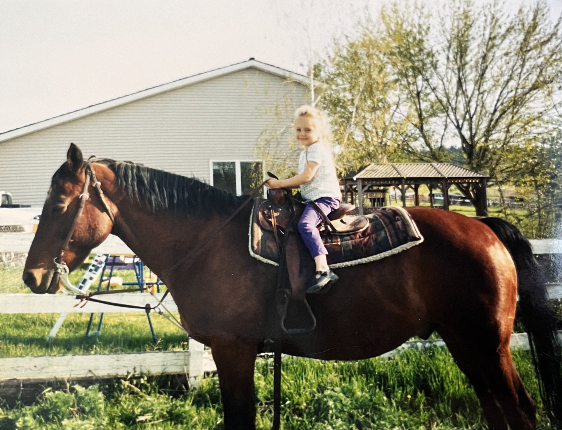 A young girl with blond hair riding a brown horse that is saddled with a patterned saddle and reins, outdoors on grass with trees and a building in the background.