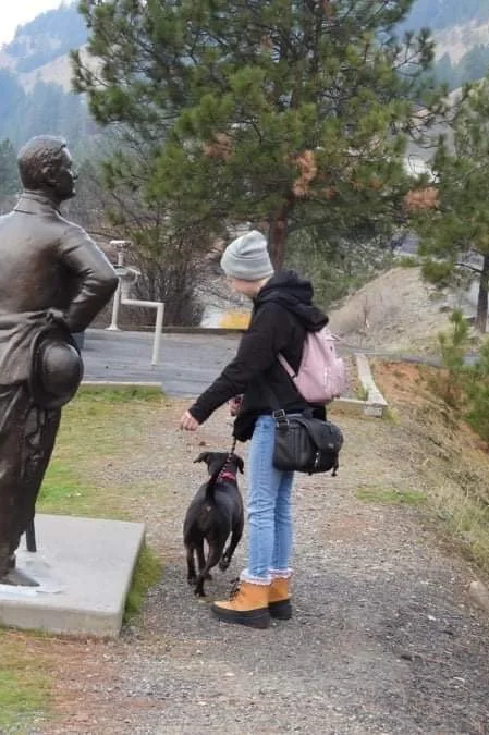 A person with a gray beanie, black jacket, blue jeans, and tan boots walks a black dog near a statue in a park with trees and mountains in the background.