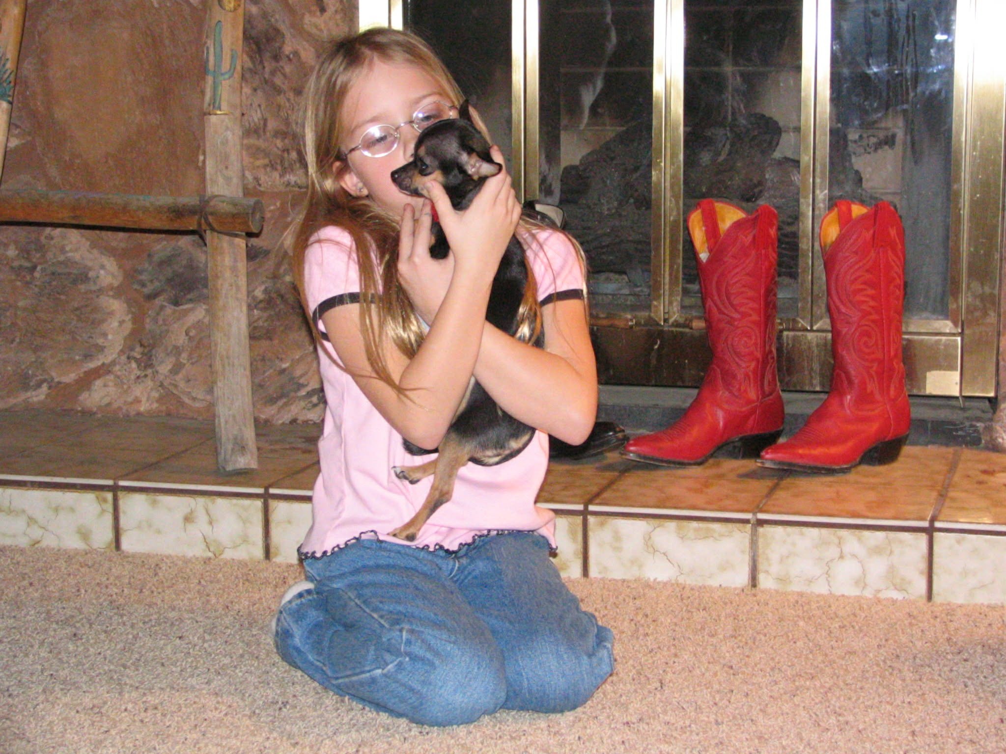 A young girl with long blonde hair, glasses, and wearing a pink shirt and blue jeans, sitting on the carpet and holding a small black puppy close to her face inside a cozy indoor space with a stone fireplace and red cowboy boots in the background.