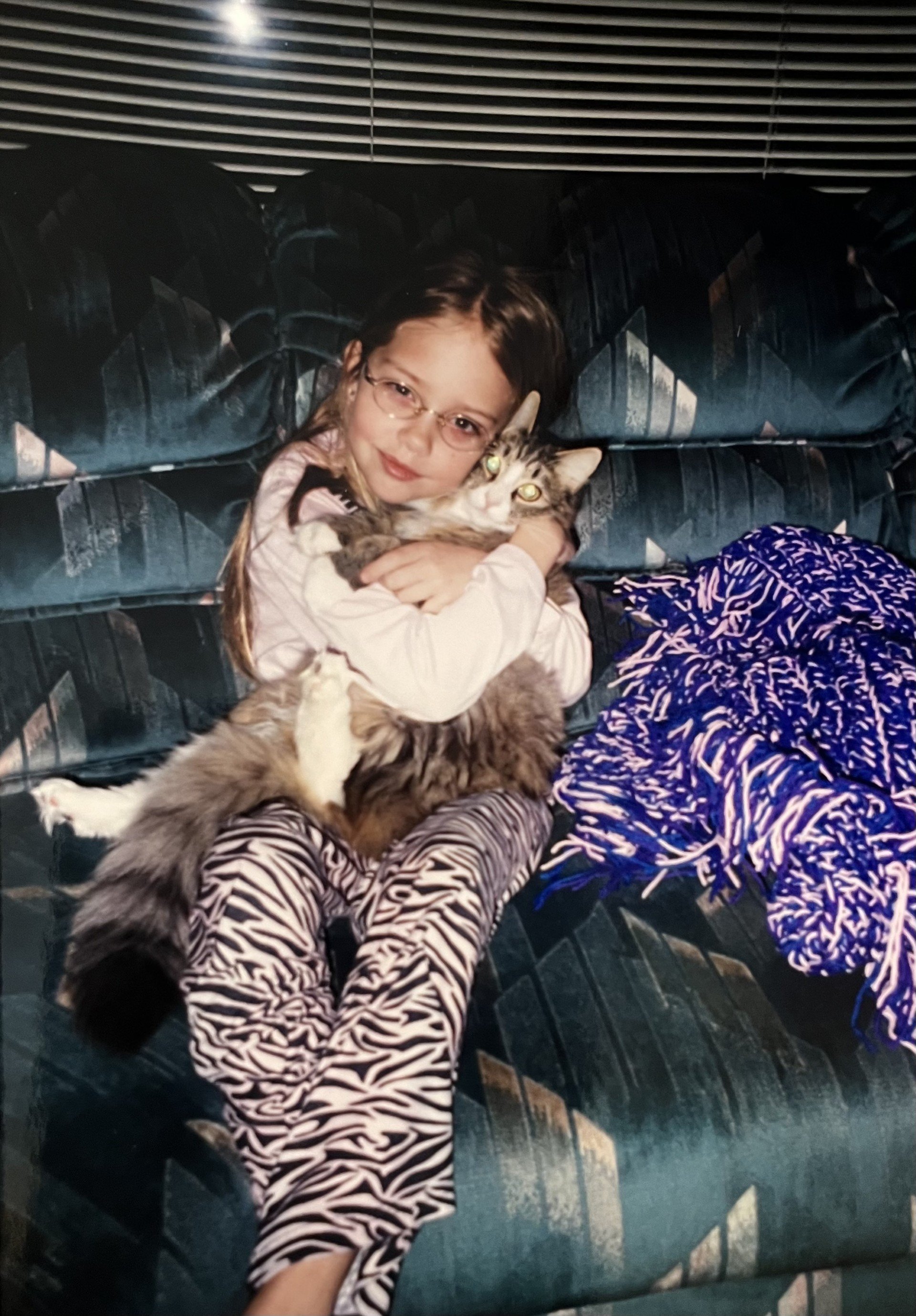 A young girl wearing glasses and zebra print pants sitting on a vintage-style dark patterned bench, hugging a long-haired tabby cat.