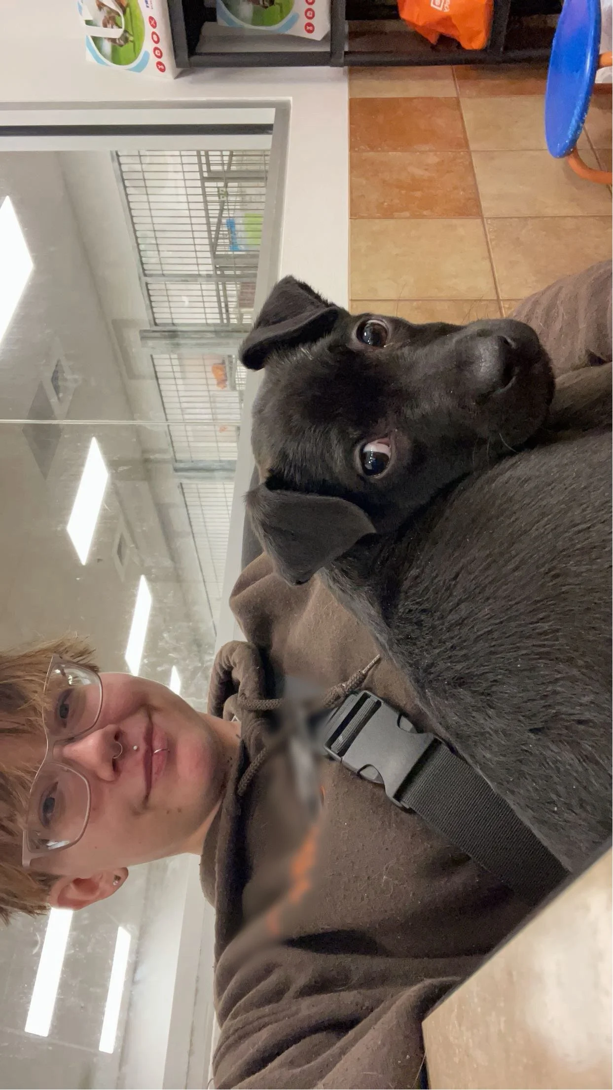 A small black puppy with brown markings is resting on a person's lap. The person has glasses and red hair. The background shows a room with tiled floors, shelves, and cages, likely a pet shelter or veterinary clinic.