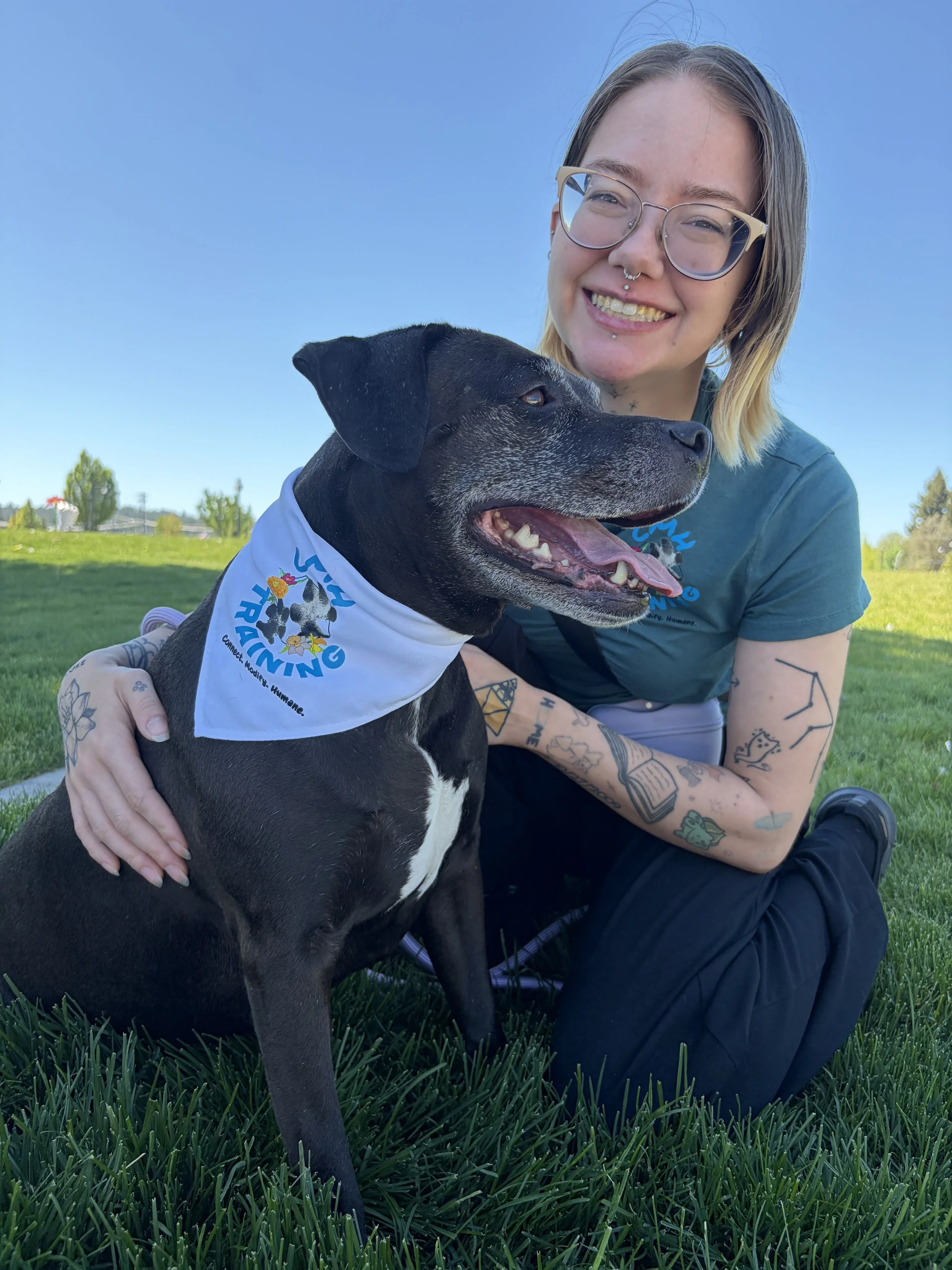 A smiling young woman with glasses and tattoos on her arms is sitting on grass outdoors with a black dog wearing a bandana. The woman has shoulder-length hair and is wearing a teal shirt. The dog appears happy with its tongue out.