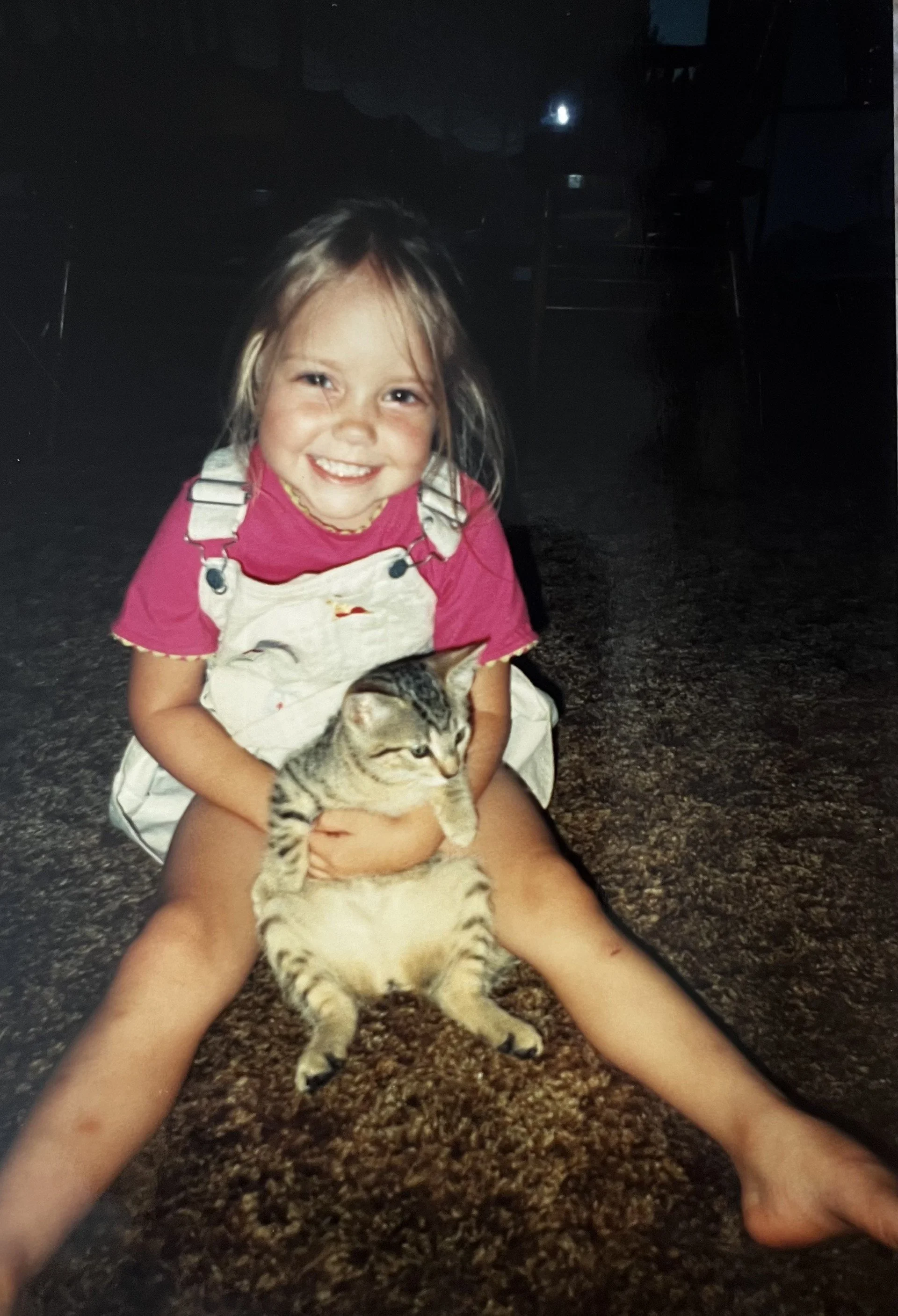 A young girl with blonde hair, wearing a pink shirt and beige overalls, is sitting on the floor holding a tabby kitten with gray and black striped fur. The girl is smiling, and the background is dark with some furniture faintly visible.