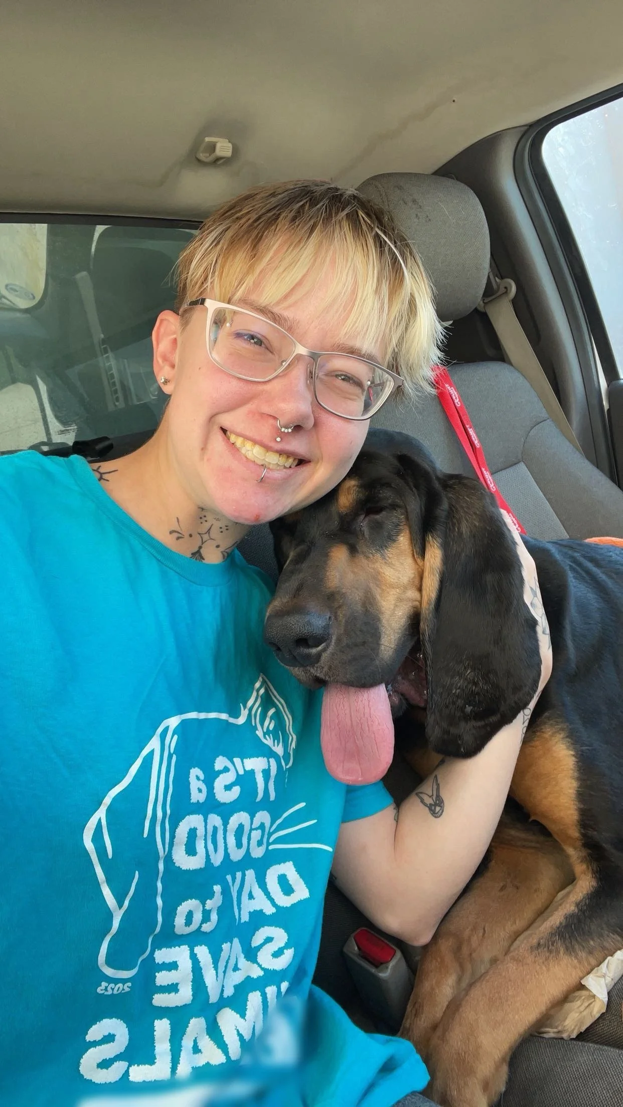 Person with short blond hair and glasses smiling while sitting in a vehicle with a large dog, possibly a bloodhound, resting on their lap. The dog has its tongue out and appears to be happy.