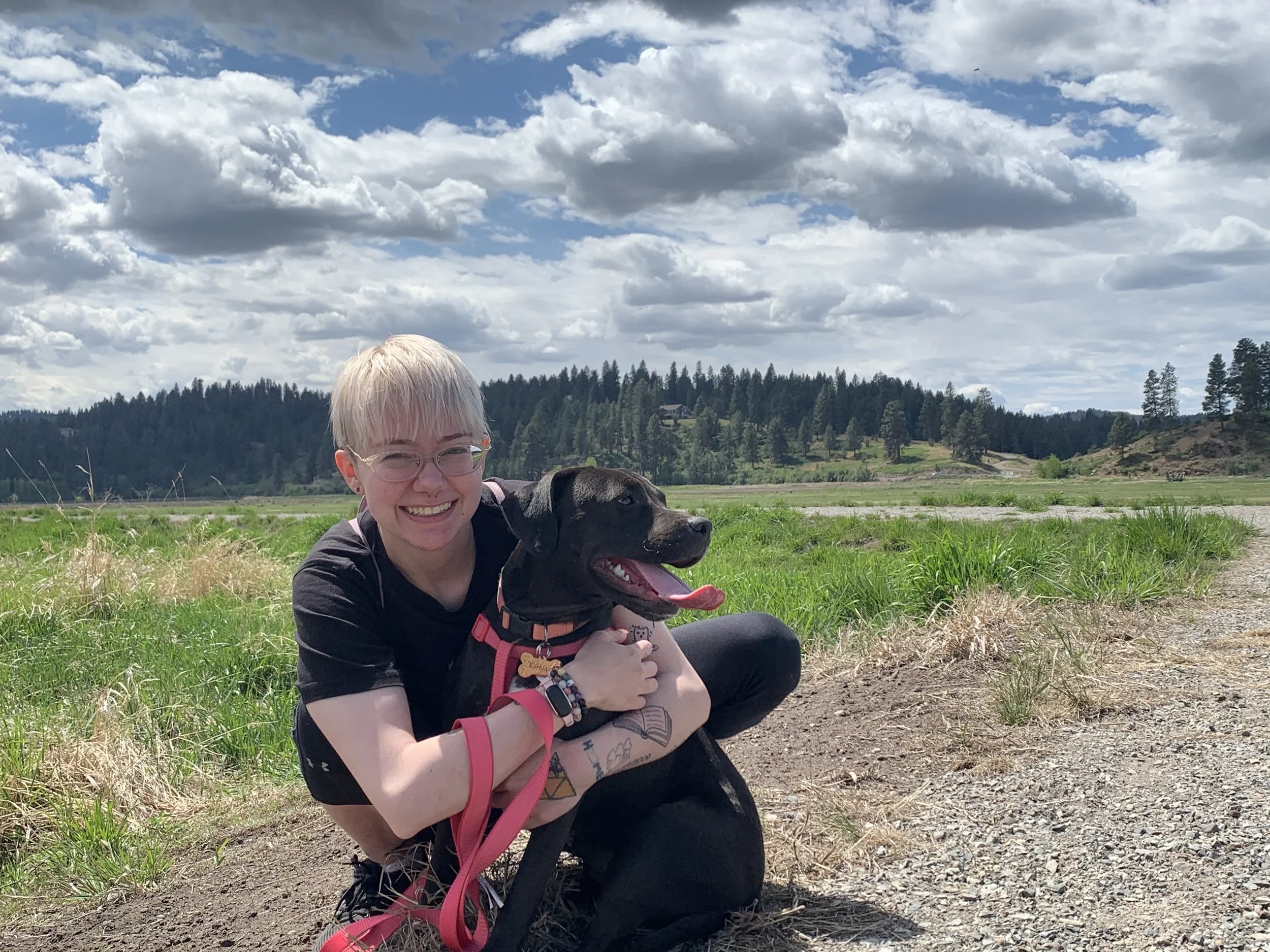 A young woman with short blonde hair, glasses, and wearing black athletic clothing, is sitting on the grass outdoors, smiling and hugging a black dog with a brown collar and a tag. Behind them are green fields, a distance of trees and hills, and a partly cloudy sky.