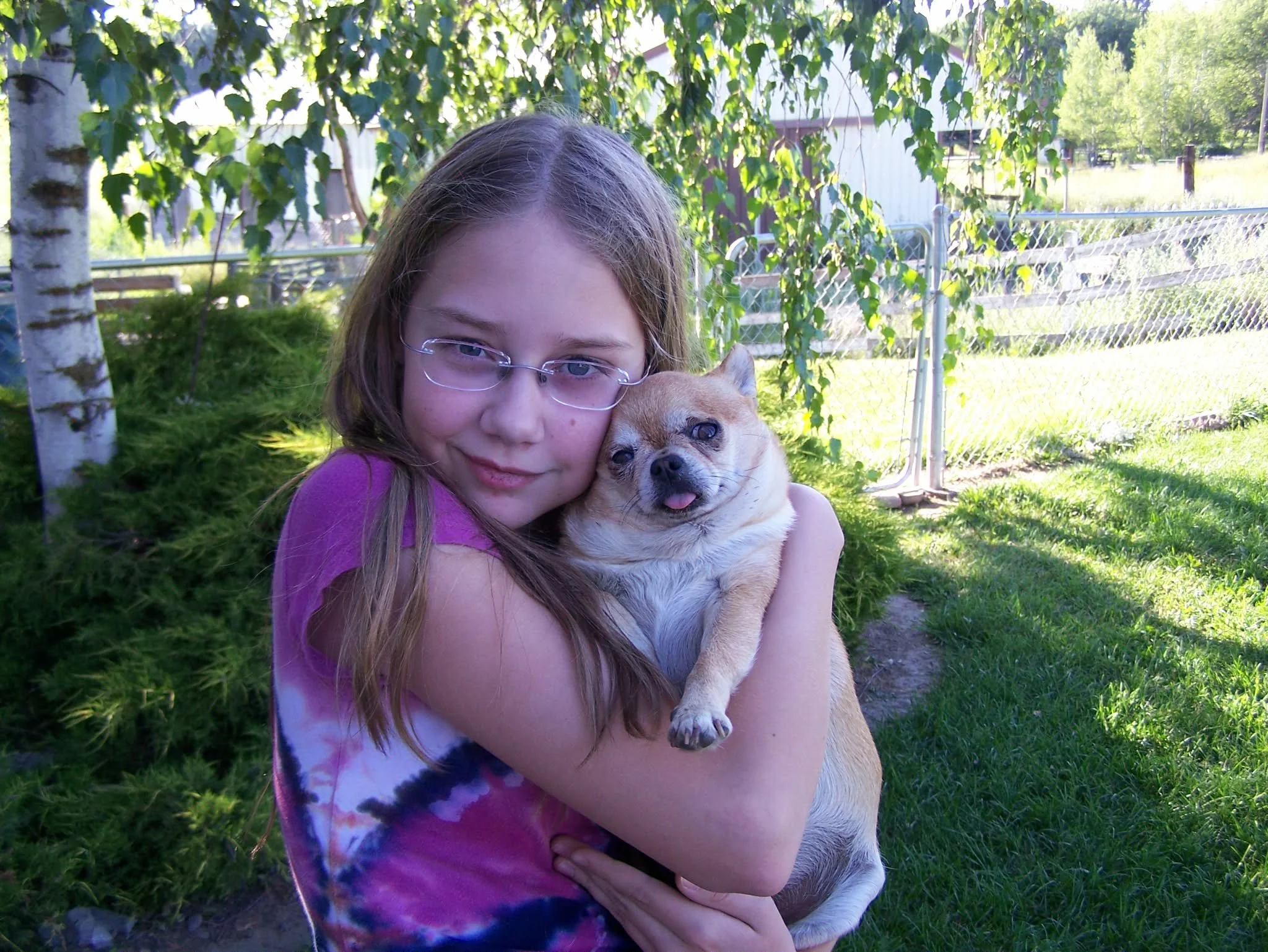 A young girl with glasses, wearing a pink and purple tie-dye shirt, hugging a small tan and white dog outdoors in a garden with trees, a fence, and a house in the background.