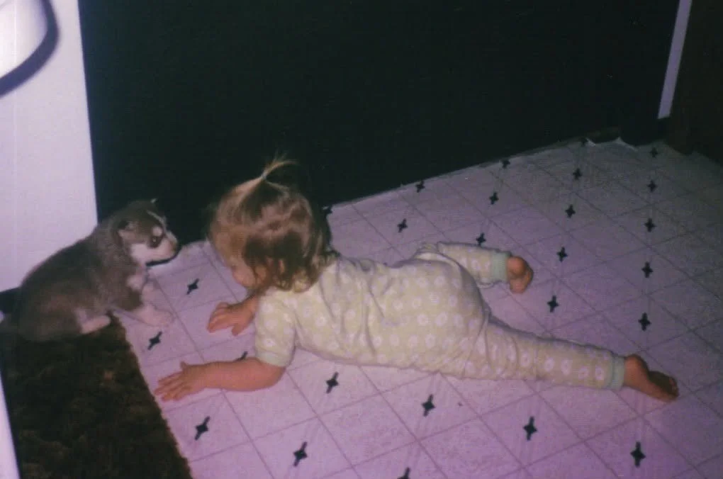 A young girl lying on her stomach on a tiled kitchen floor, facing a raccoon sitting upright near her.