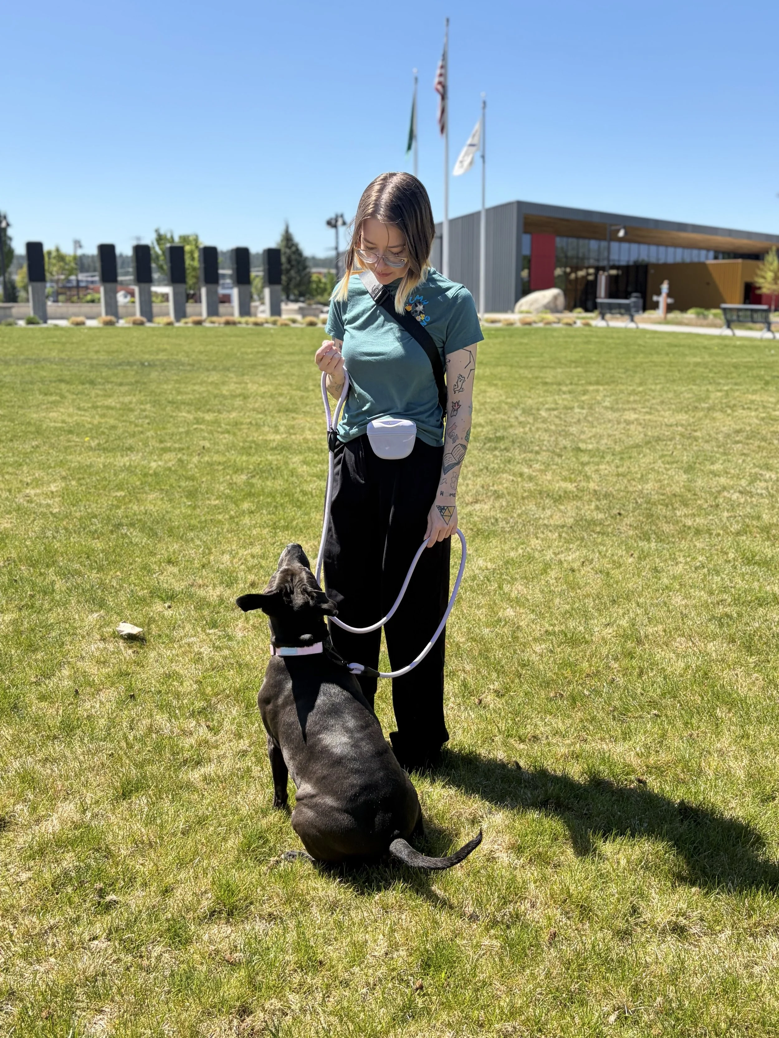 A young woman with tattoos on her arms, wearing glasses, a turquoise t-shirt, and black pants, is standing outdoors on a sunny day with a small black and white dog on a leash. She is looking down at the dog, who is sitting on the grass, with a modern building and flags in the background.