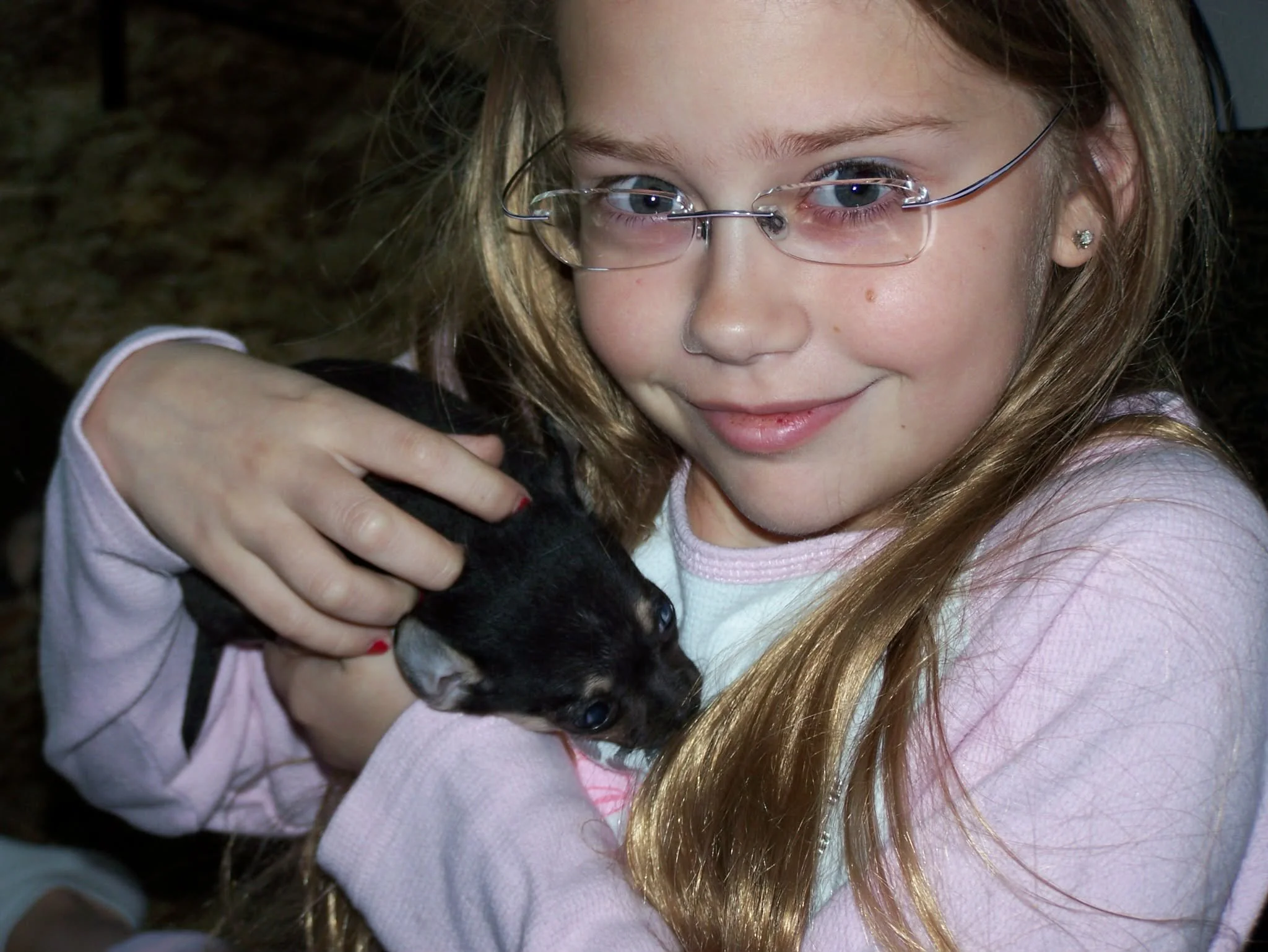 A young girl with long blonde hair and glasses holding a black puppy.