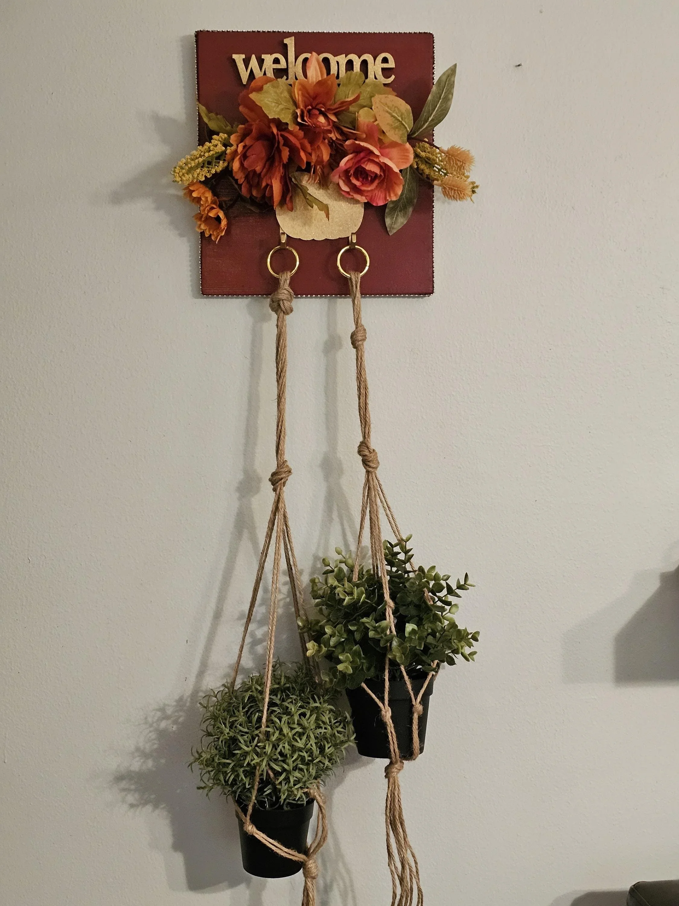 A decorative wall hanging with a red square background that says 'welcome' at the top, adorned with artificial orange and pink flowers, green leaves, and a small beige circle with two hanging hooks. Below it, two small black pots with green plants hang from beige ropes.