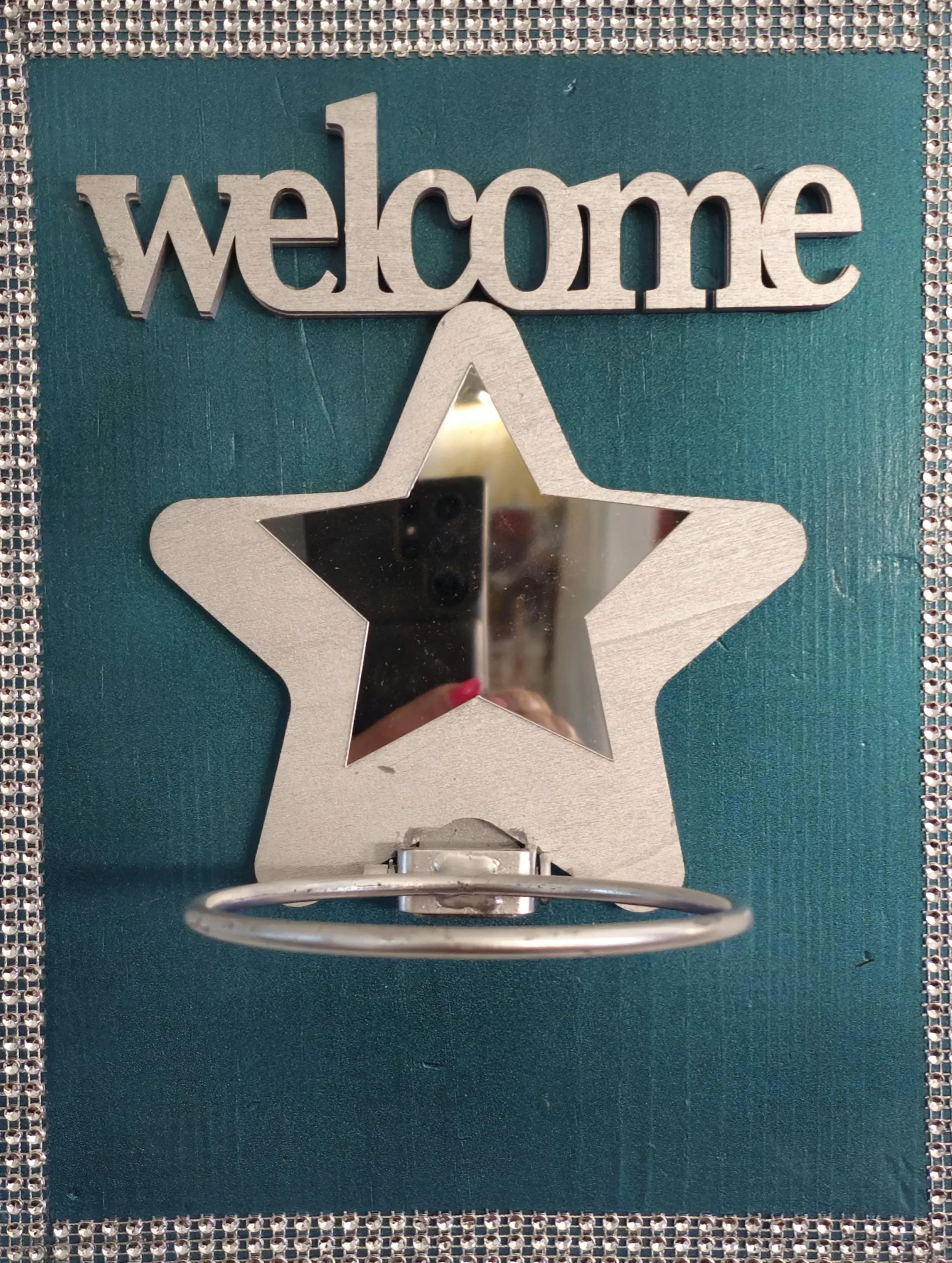 Decorative wall arrangement with a silver 'welcome' sign and a star-shaped mirror on teal background with a rhinestone border.