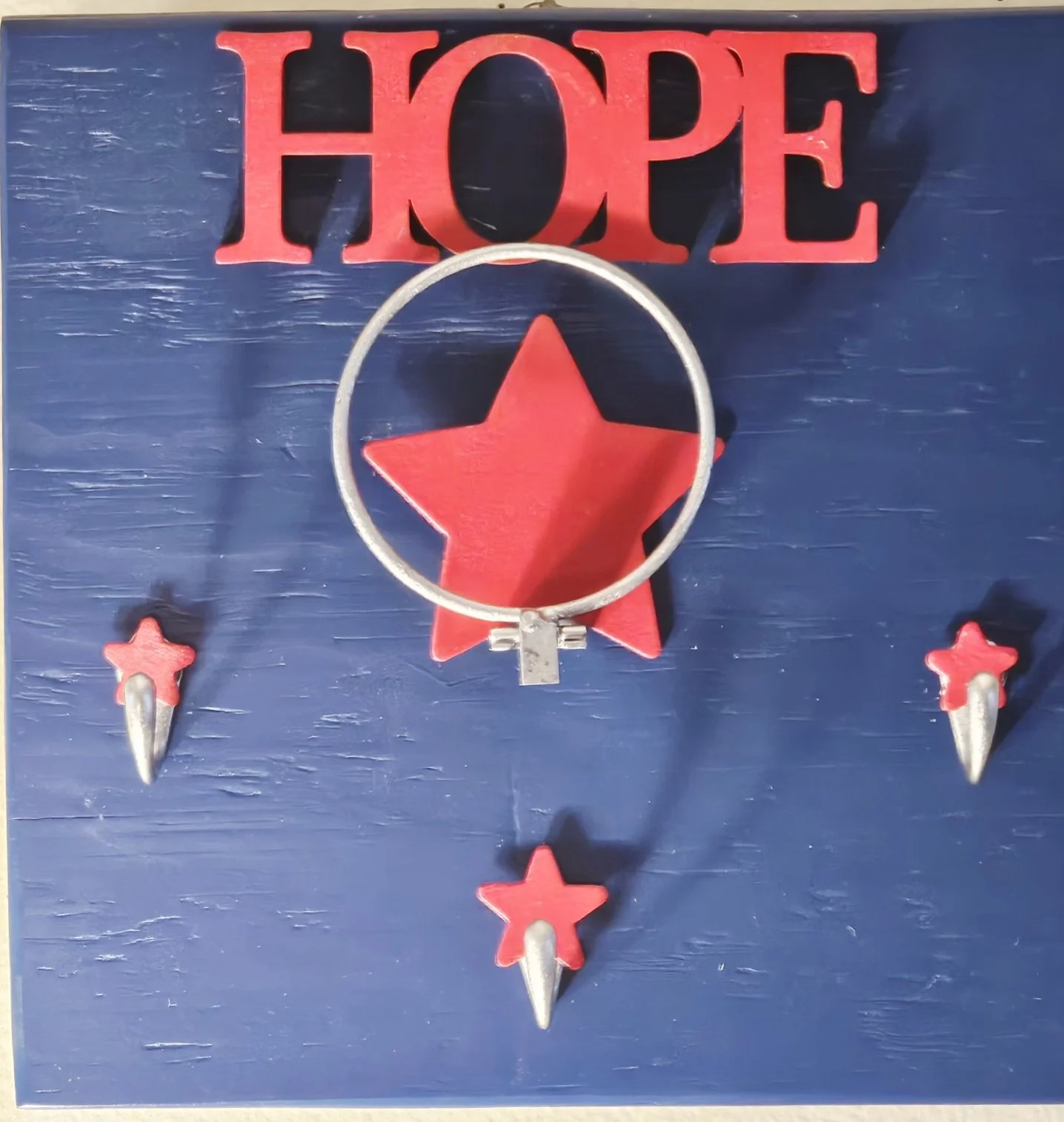 Decorative wall art with the word 'HOPE' in red, a red star inside a silver ring, and four smaller red stars with silver points on a blue wooden background.