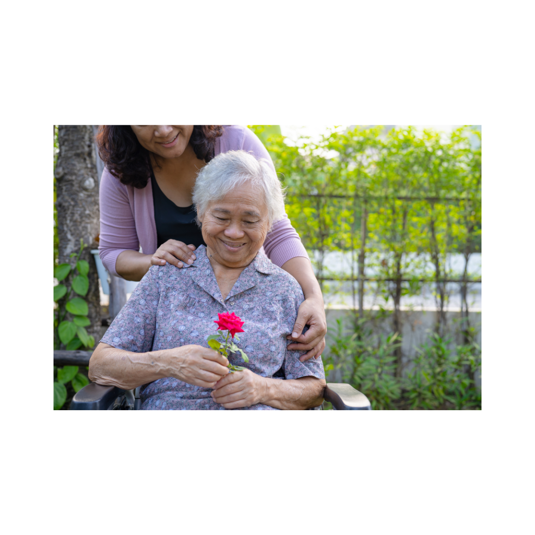 An elderly woman with white hair smiling and holding a pink flower in her hands, with a younger woman standing behind her outdoors in a garden.