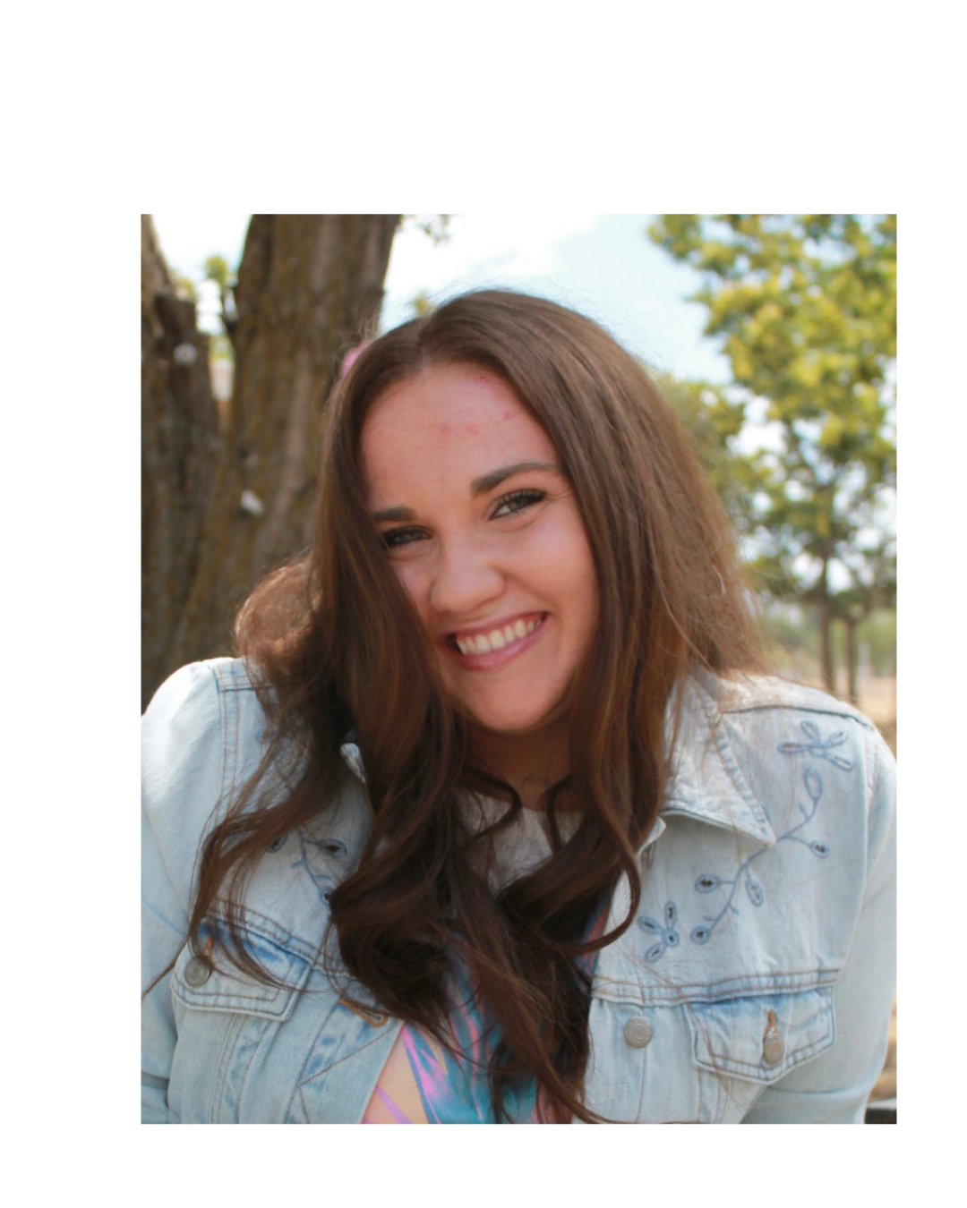 Smiling young woman with long brown hair, wearing a light denim jacket with floral embroidery, outdoors in a park with trees and blue sky.