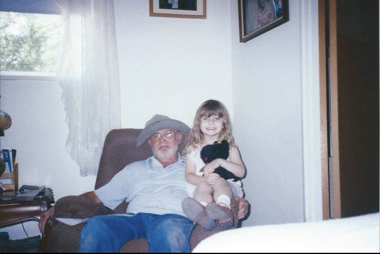 A man sitting in a brown armchair, wearing glasses, a light blue shirt, and a gray hat, with a young girl sitting on his lap holding a black stuffed animal, smiling. There is a window with white curtains behind them and framed pictures on the wall.