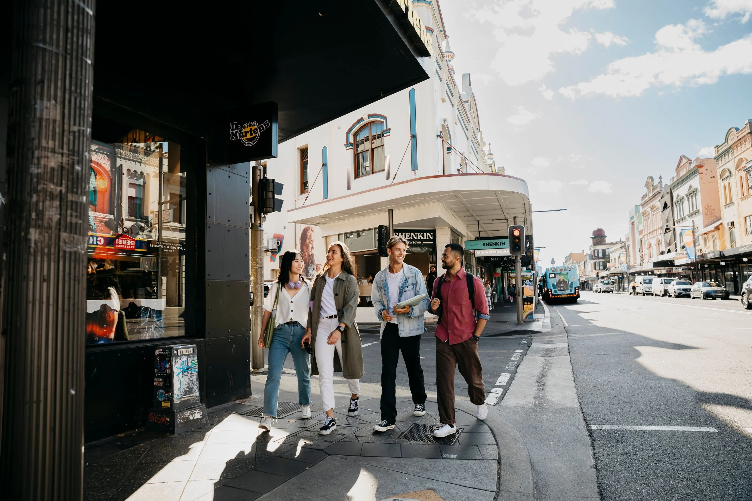 A group of five young people walking together on a city sidewalk, talking and smiling. They are near storefronts and a bus is visible on the busy street. The sky is partly cloudy and the scene is brightly lit.