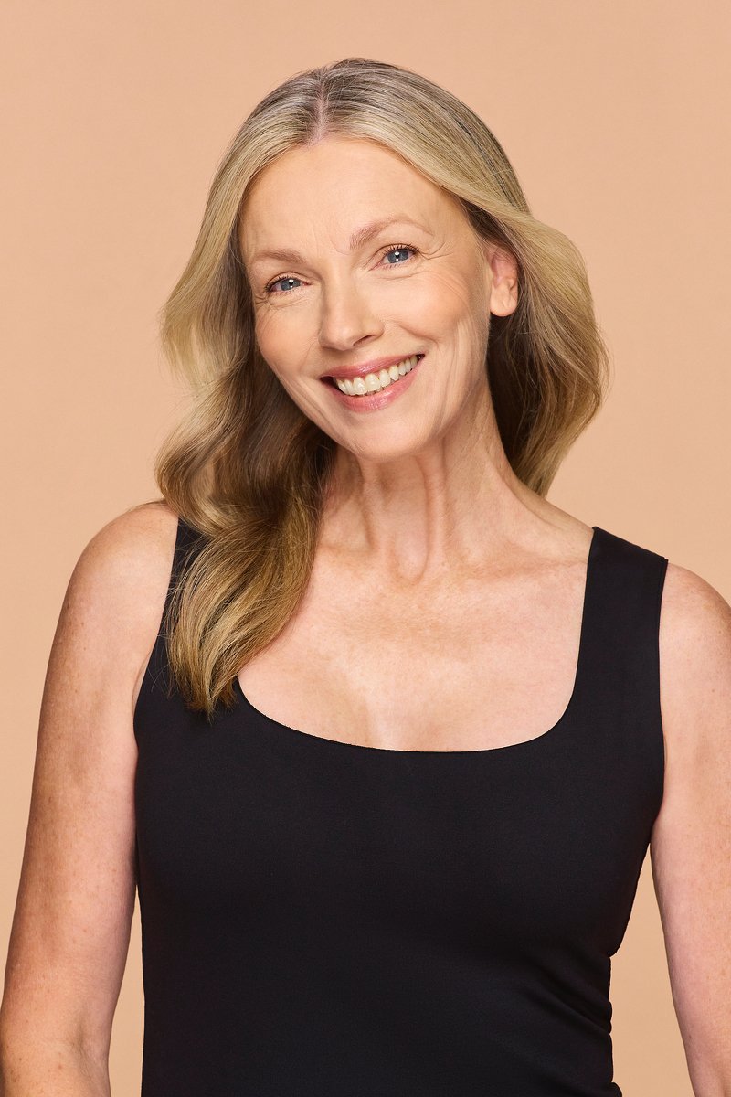 Portrait of a smiling middle-aged woman with blonde hair wearing a black sleeveless top, against a beige background.