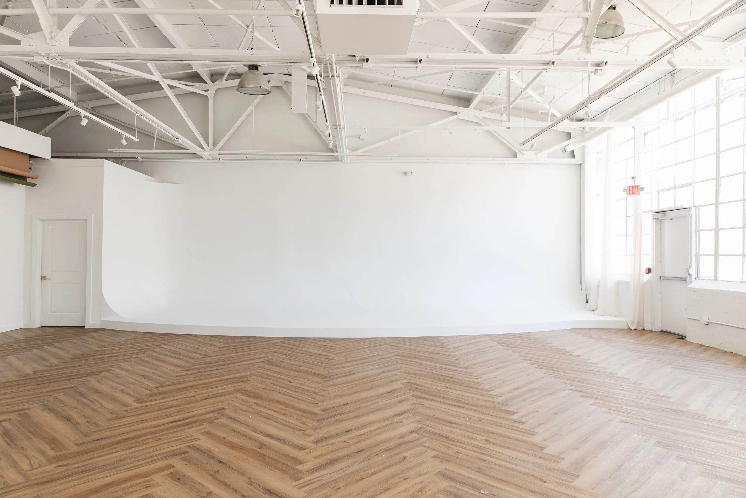 Empty room with white walls, large windows, and a wooden floor arranged in a herringbone pattern, with exposed ceiling beams and industrial lighting.