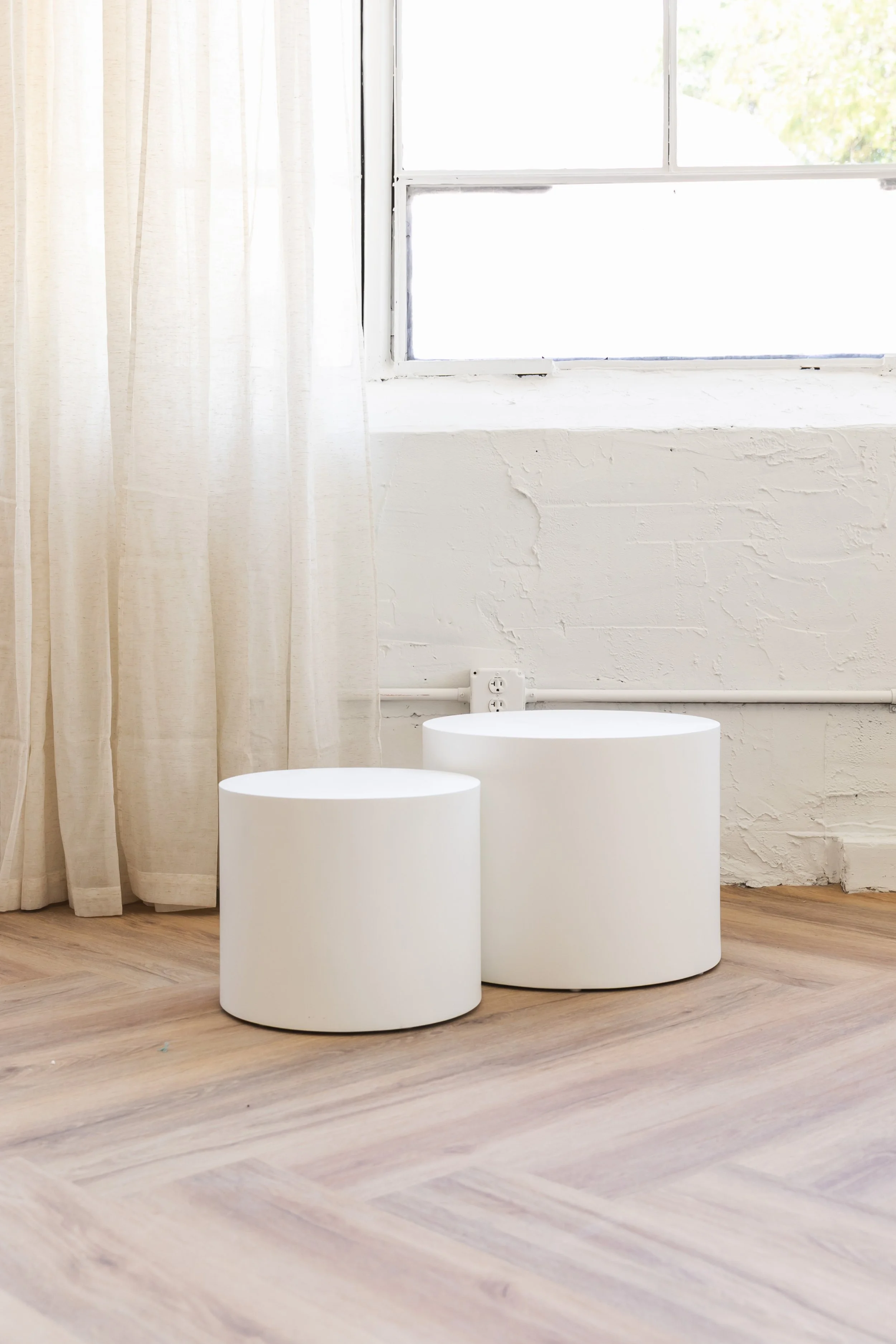 Two minimalist white cylindrical stools are placed on a wooden floor in front of a white wall near a window with beige curtains.