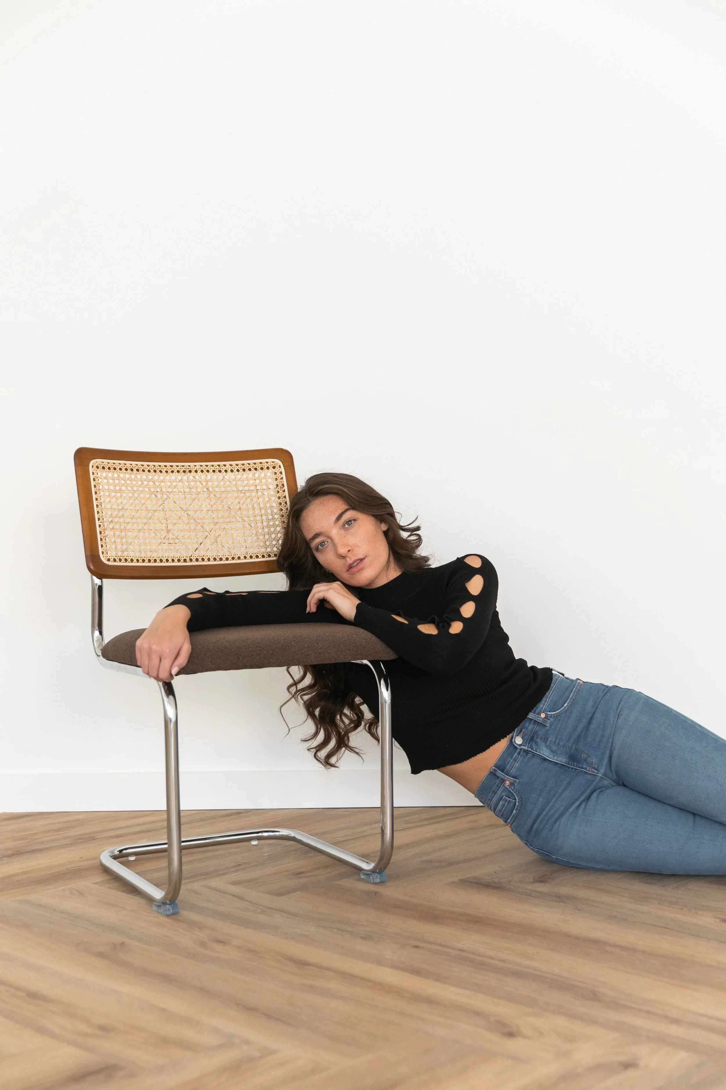 A woman lying on the wooden floor with her head resting on a vintage chair, wearing a black sweater with cutouts on the sleeves and blue jeans, against a plain white wall.