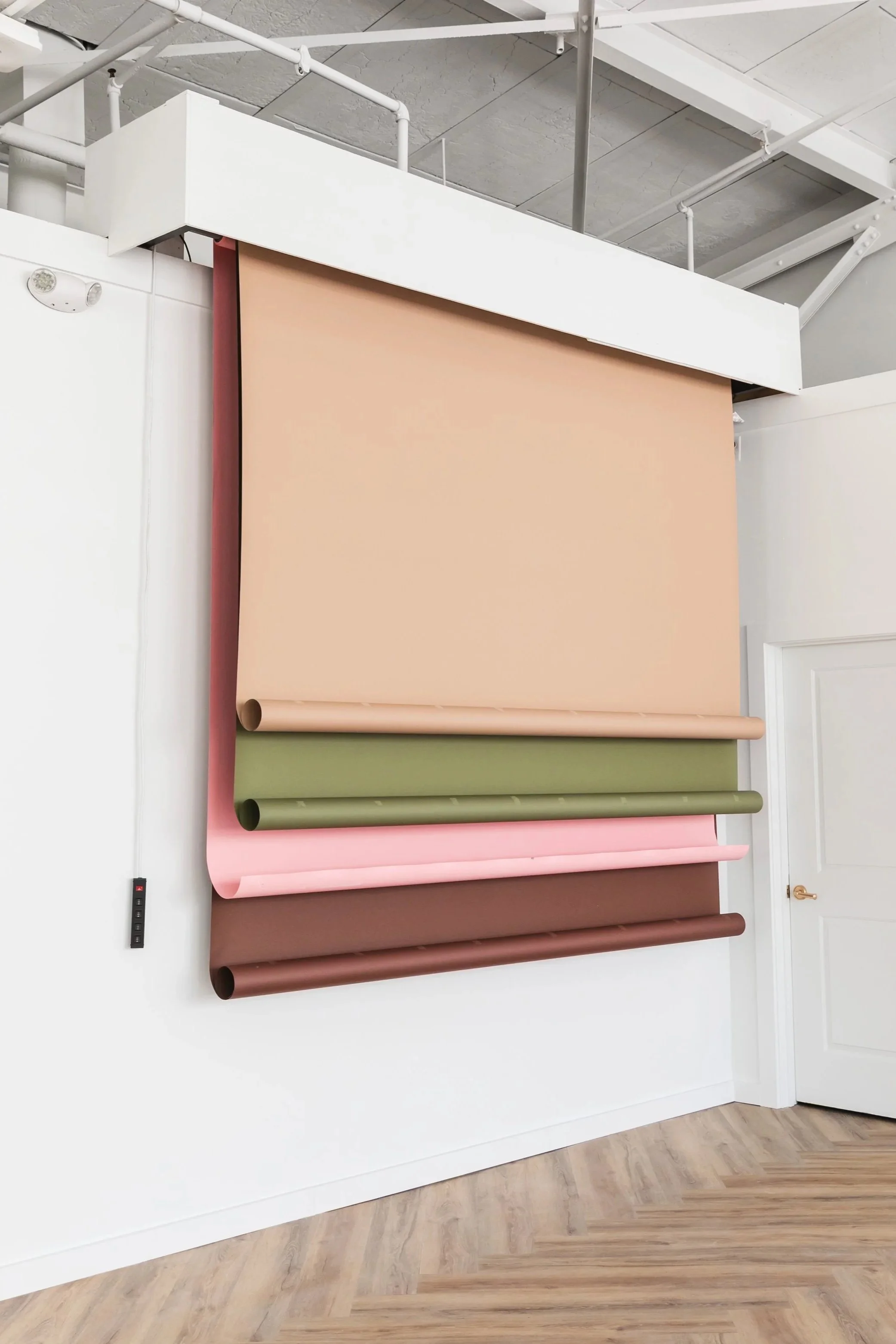 Multiple rolls of wallpaper hanging vertically on a white wall in an interior room with wooden flooring.