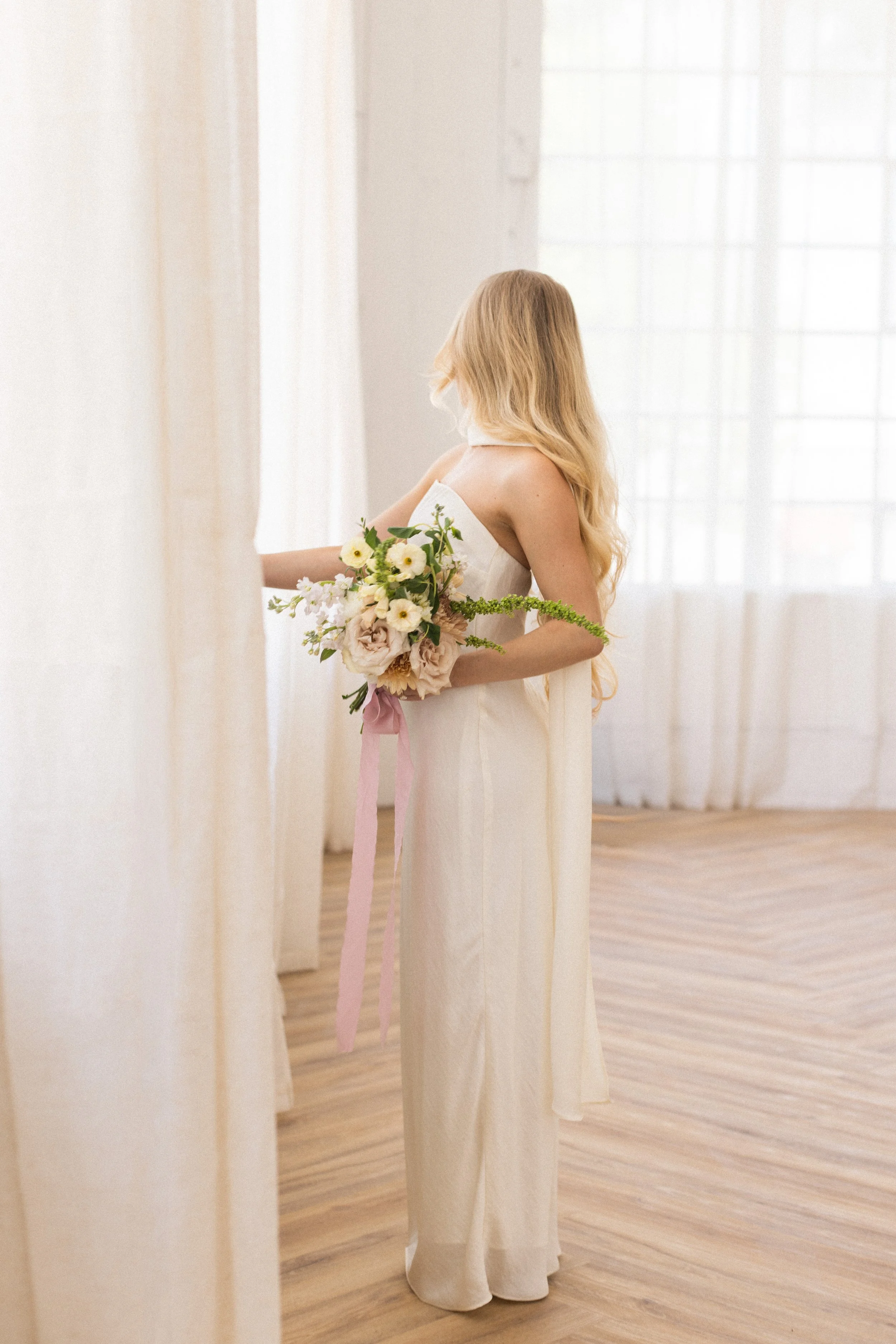 A woman in a white dress holding a bouquet of white and pink flowers, standing in a softly lit room with sheer curtains and wooden floor.